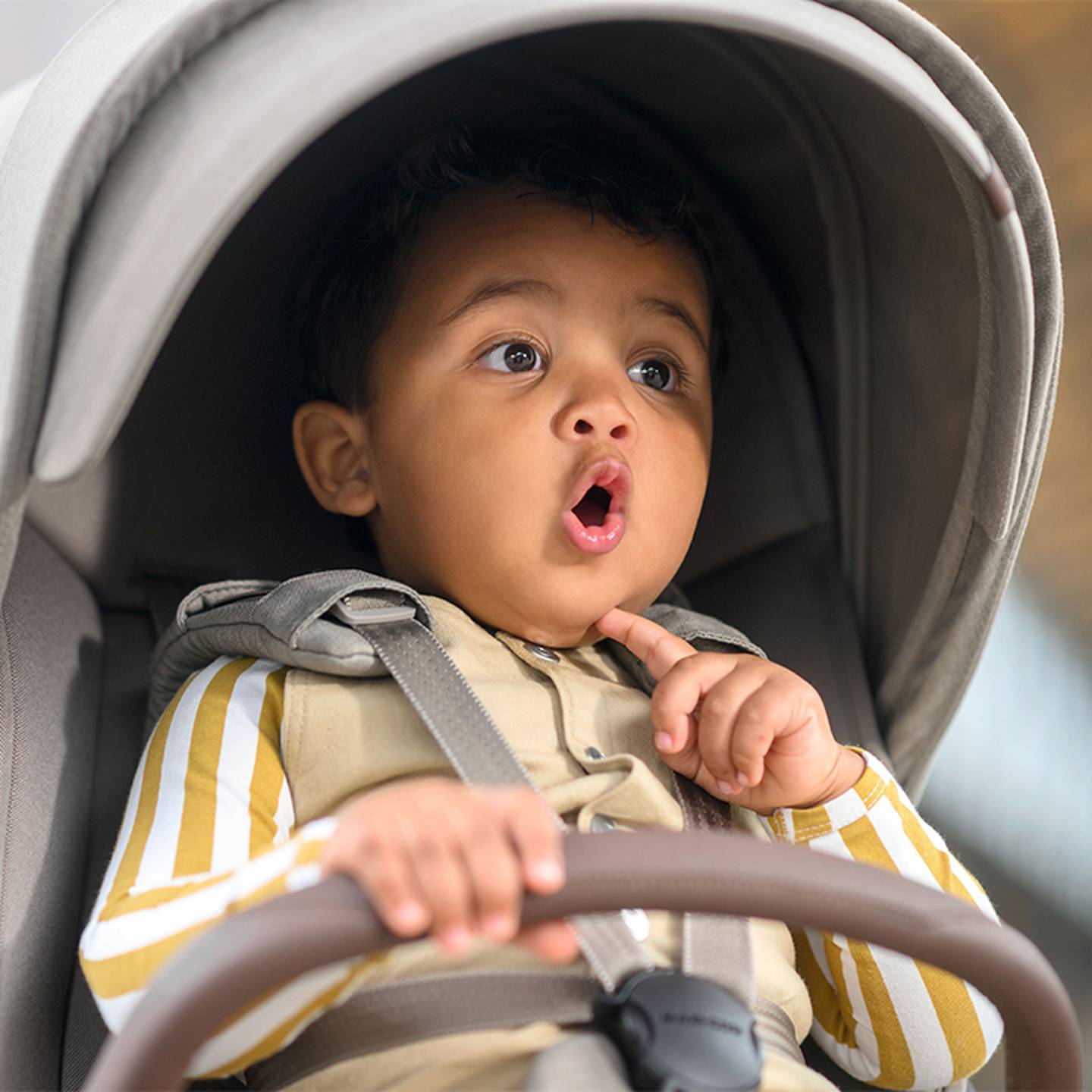 Kleinkind im Kinderwagen, mit gestreiftem Hemd, überraschtem Gesichtsausdruck und offenem Mund, die Hand ruht am Kinn.