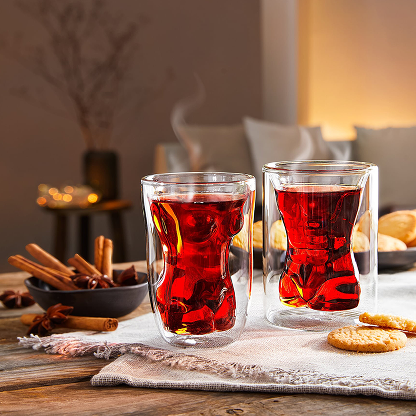 Zwei Gläser dampfender roter Glühwein auf einem Holztisch, umgeben von Zimtstangen und Keksen, die für eine gemütliche Atmosphäre sorgen.