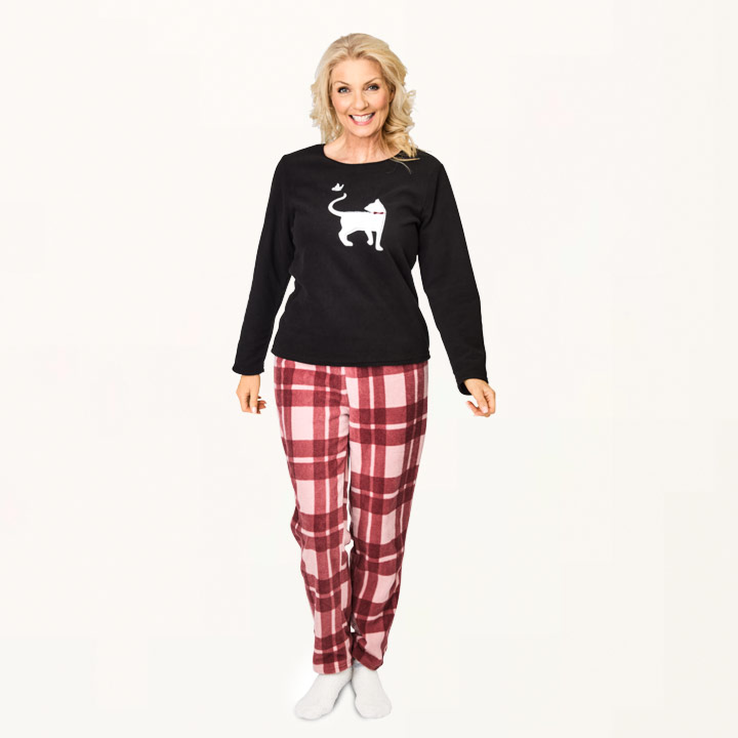Frau in einem schwarzen Pullover mit Katzenmotiv und einer rotkarierten Pyjamahose, lächelnd vor weißem Hintergrund.
