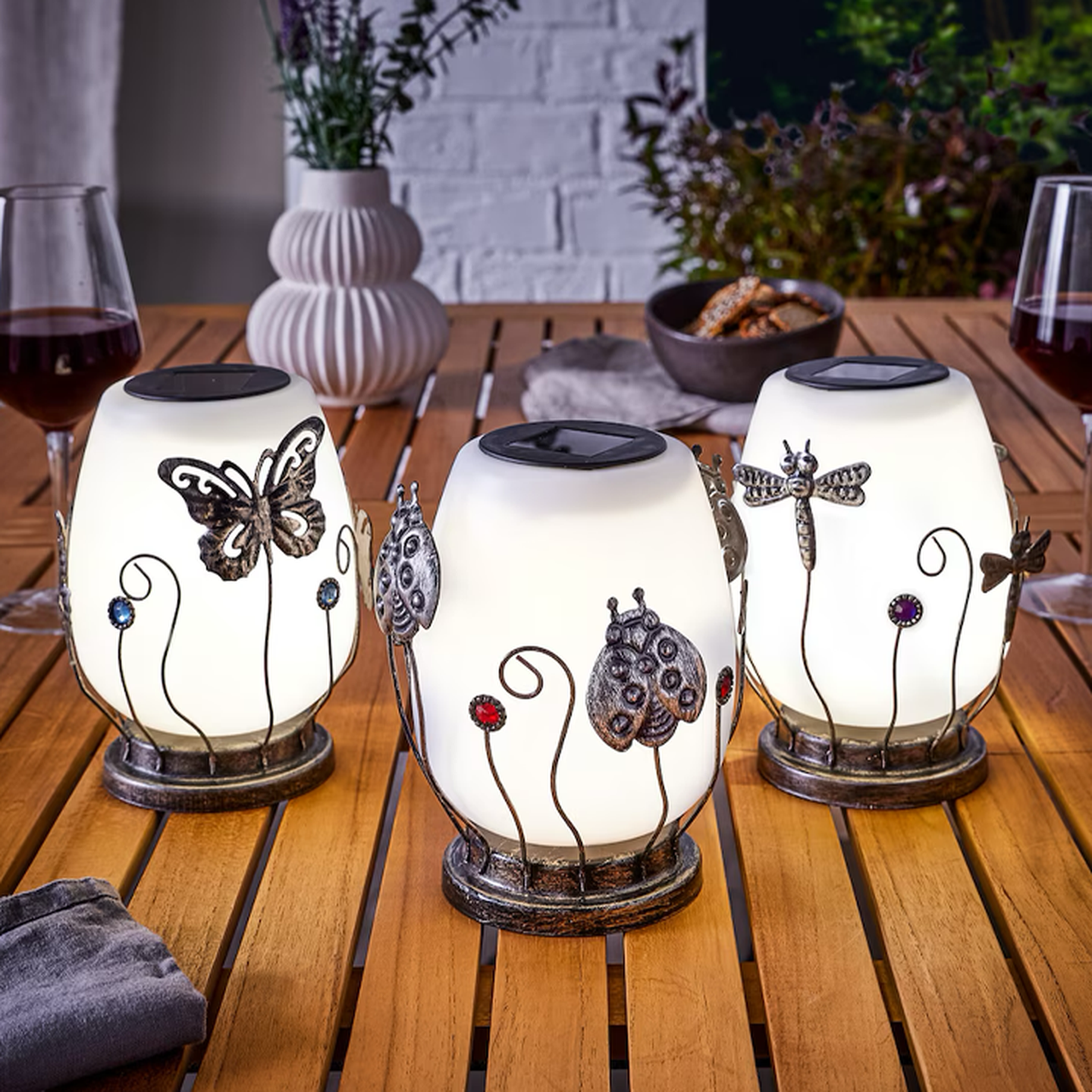 Drei beleuchtete, dekorative Laternen mit Insektenmotiven auf einem Holztisch, umgeben von Rotweingläsern und einer Pflanzenvase.
