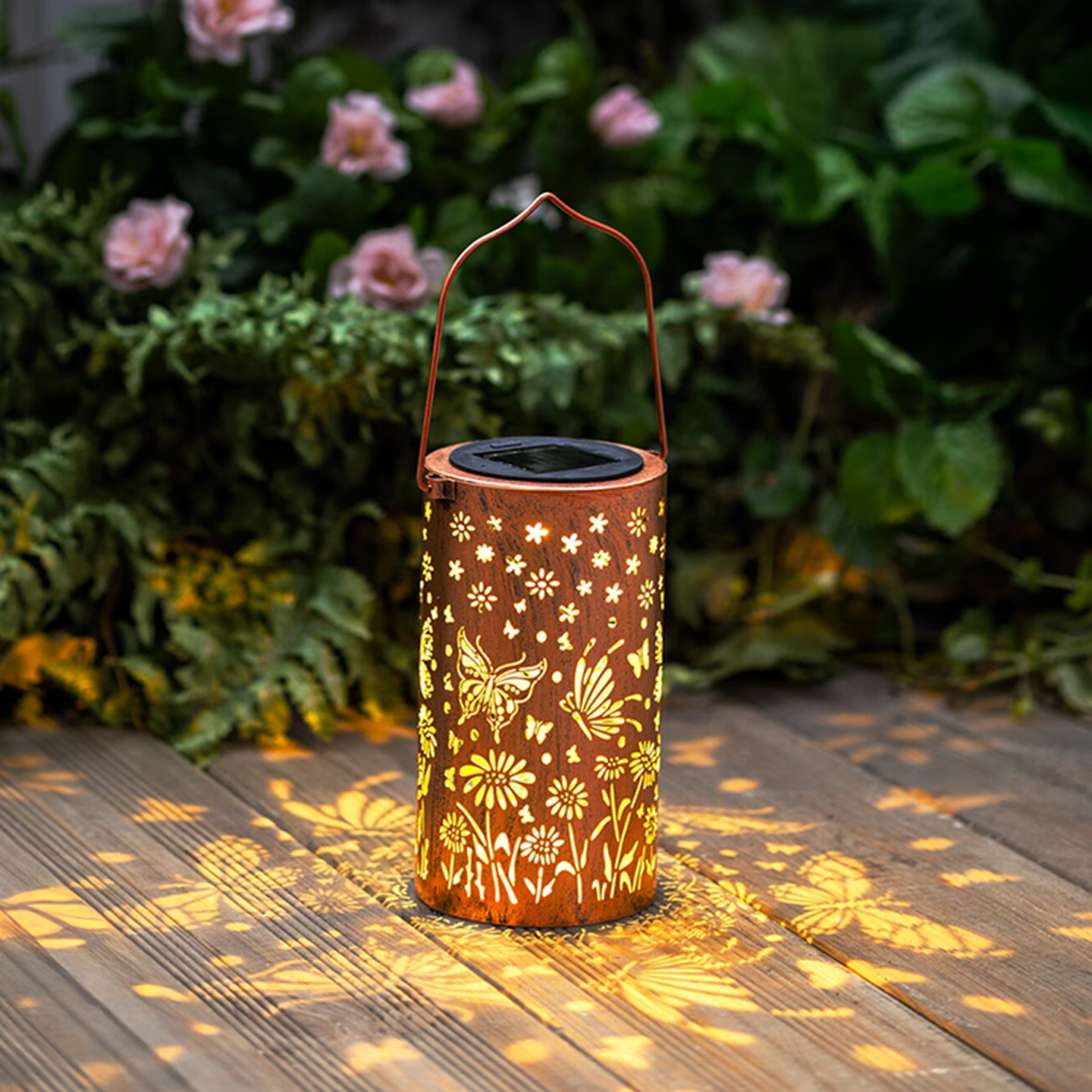 Eine dekorative Laterne mit Schmetterlings- und Blumen-Ausstanzungen, die warm leuchtende Muster auf eine Holzterrasse wirft, umgeben von sattgrünem Blattwerk und rosa Blüten.