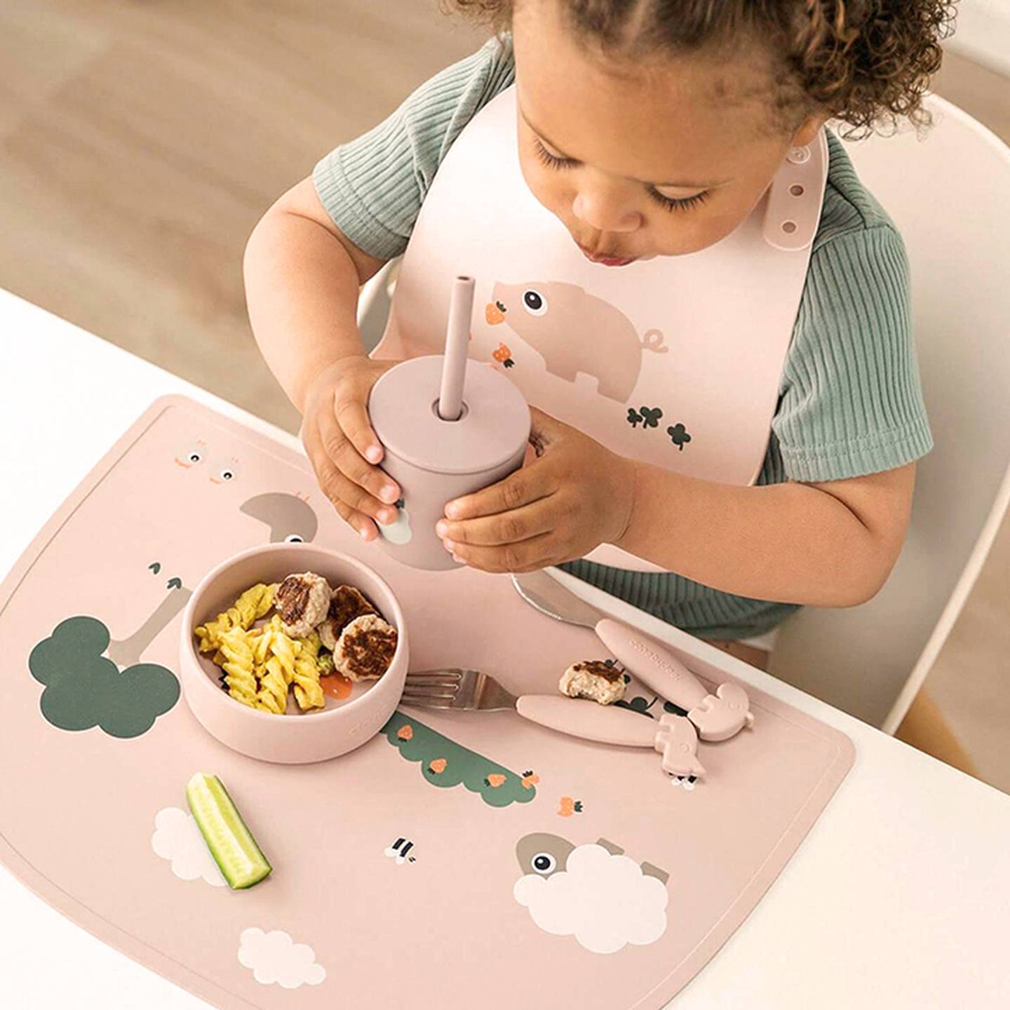 Ein Kleinkind in einem grünen Hemd und mit einem Lätzchen mit Tiermotiven sitzt an einem Tisch und isst Nudeln mit Fleischbällchen aus einem Strohhalmbecher auf einem dekorativen Untersetzer.