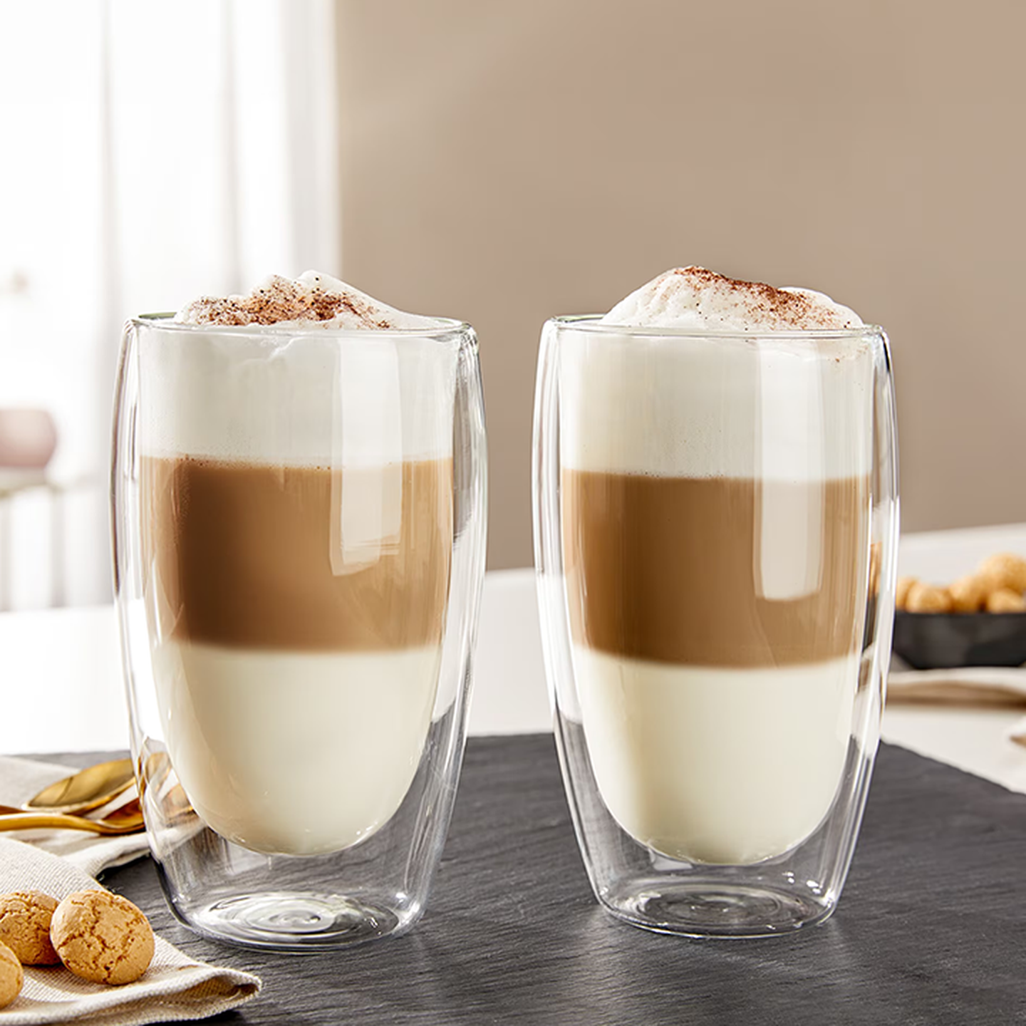 Zweischichtige Cappuccinos in durchsichtigen doppelwandigen Gläsern mit Milchschaumhaube und Kakaopulver bestreut, auf einer dunklen Oberfläche neben Keksen platziert.