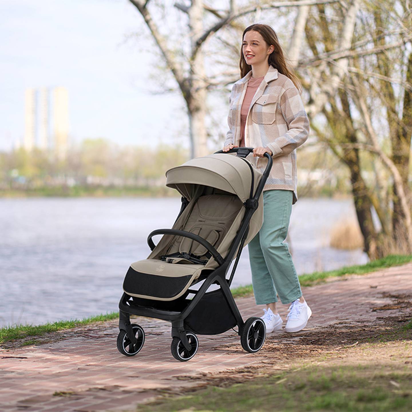 Eine Frau in Freizeitkleidung schlendert an einem sonnigen Tag mit einem leeren Kinderwagen an einem von Bäumen gesäumten Flussweg entlang.