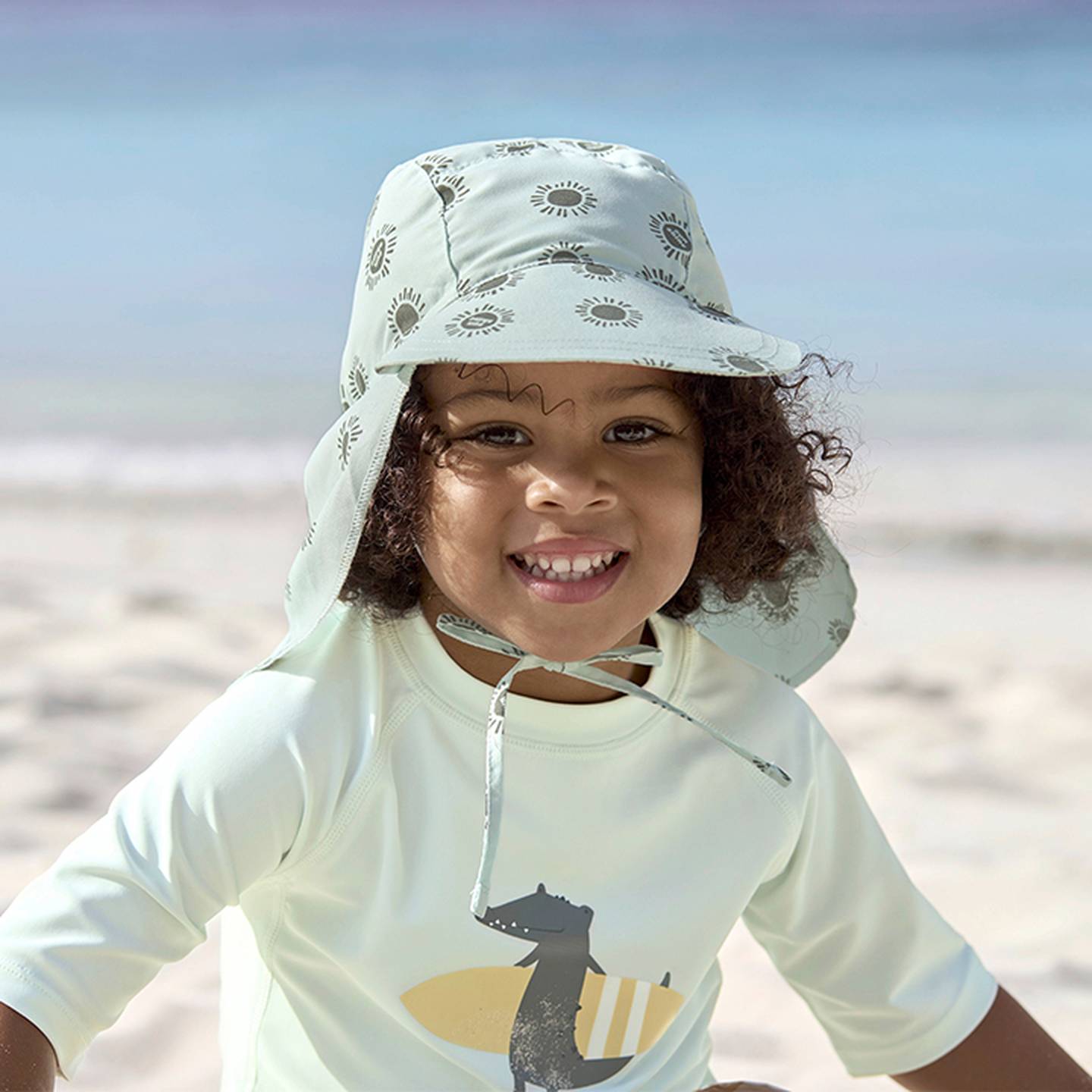 Ein lächelndes Kind mit Sonnenhut und einem Hemd mit verspieltem Muster genießt einen sonnigen Tag am Strand.