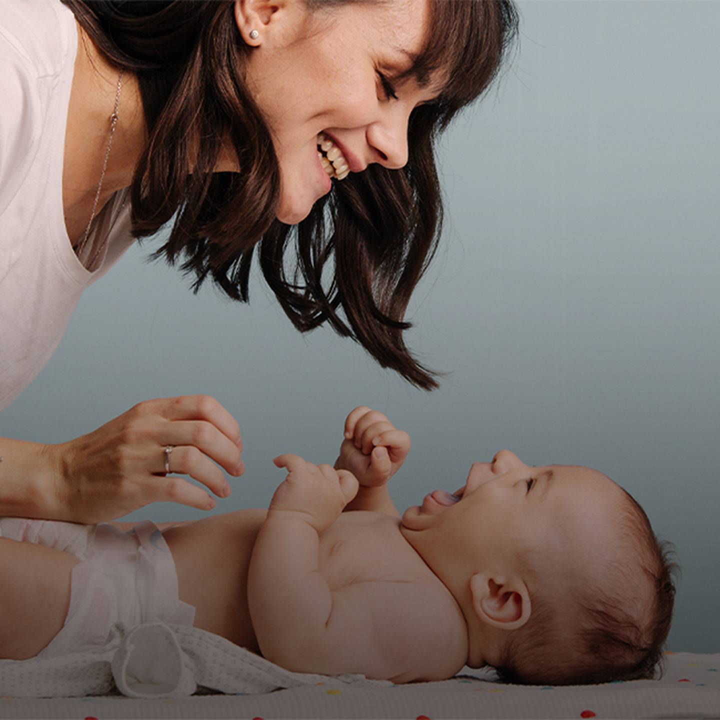 Eine Frau lächelt und spielt mit einem lachenden Baby, das vor einem hellblauen Hintergrund auf einem Wickeltisch liegt.