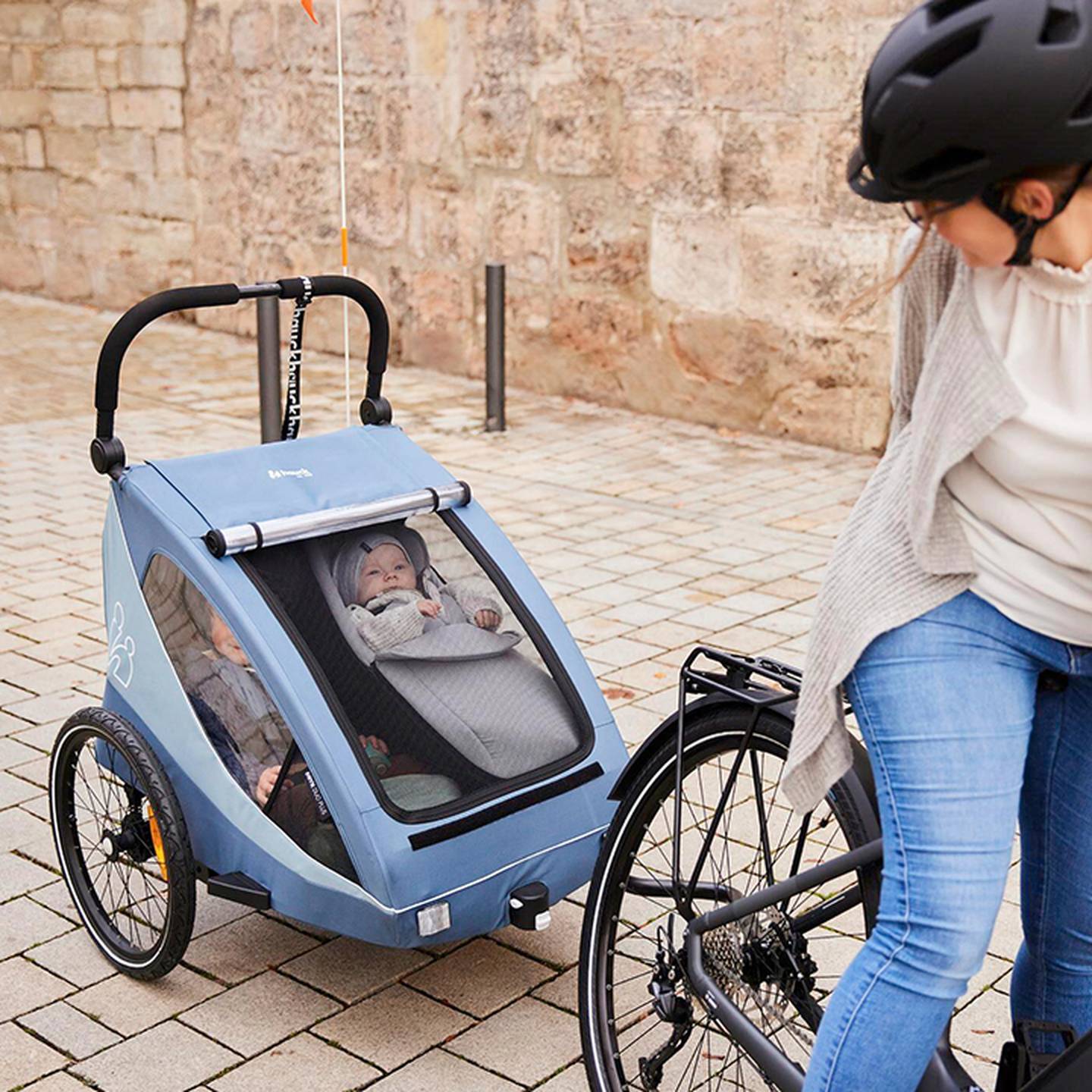 Eine Person fährt mit dem Fahrrad und hat einen blauen Kinderanhänger angehängt, in dem zwei kleine Kinder transportiert werden, von denen eines aus dem Anhänger herausschaut.