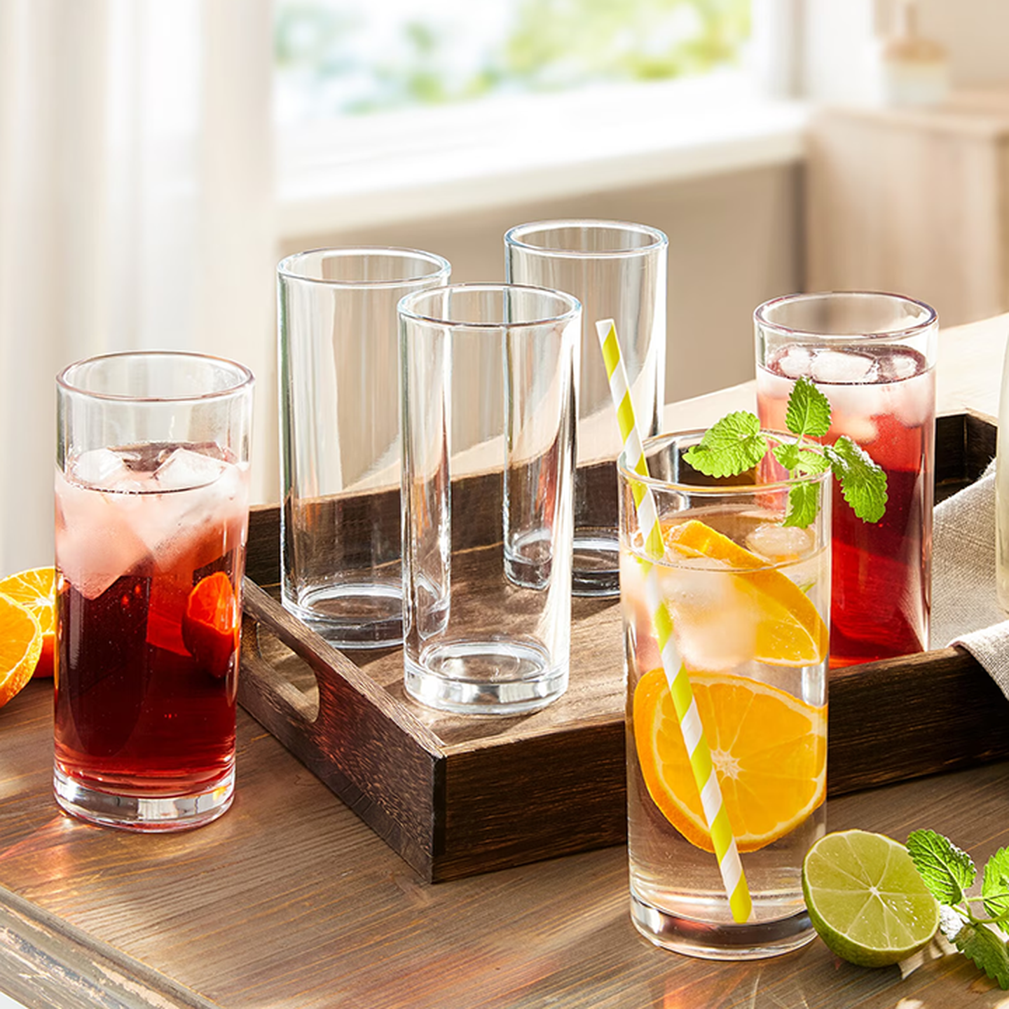 Zwei mit Eis gefüllte Gläser – eines mit roter Flüssigkeit, das andere mit Zitrusscheiben – stehen neben zwei leeren Gläsern auf einem Tablett; Zitronen und Limetten liegen daneben.