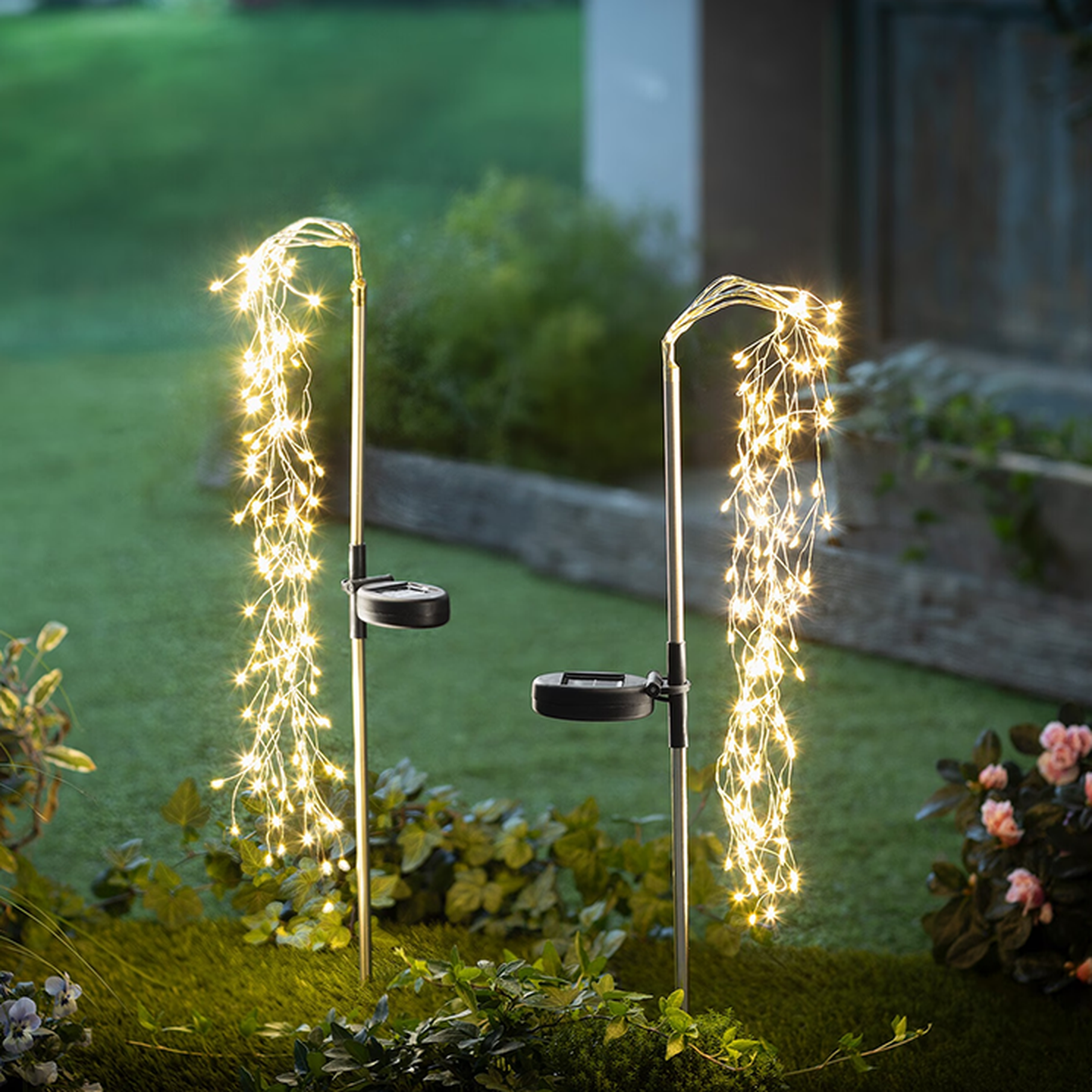 Zwei Gartenstecker, geschmückt mit kaskadierenden, warmweißen Lichterketten, eingebettet in üppiges Grün – für eine zauberhafte Atmosphäre im Freien bei Dämmerung.