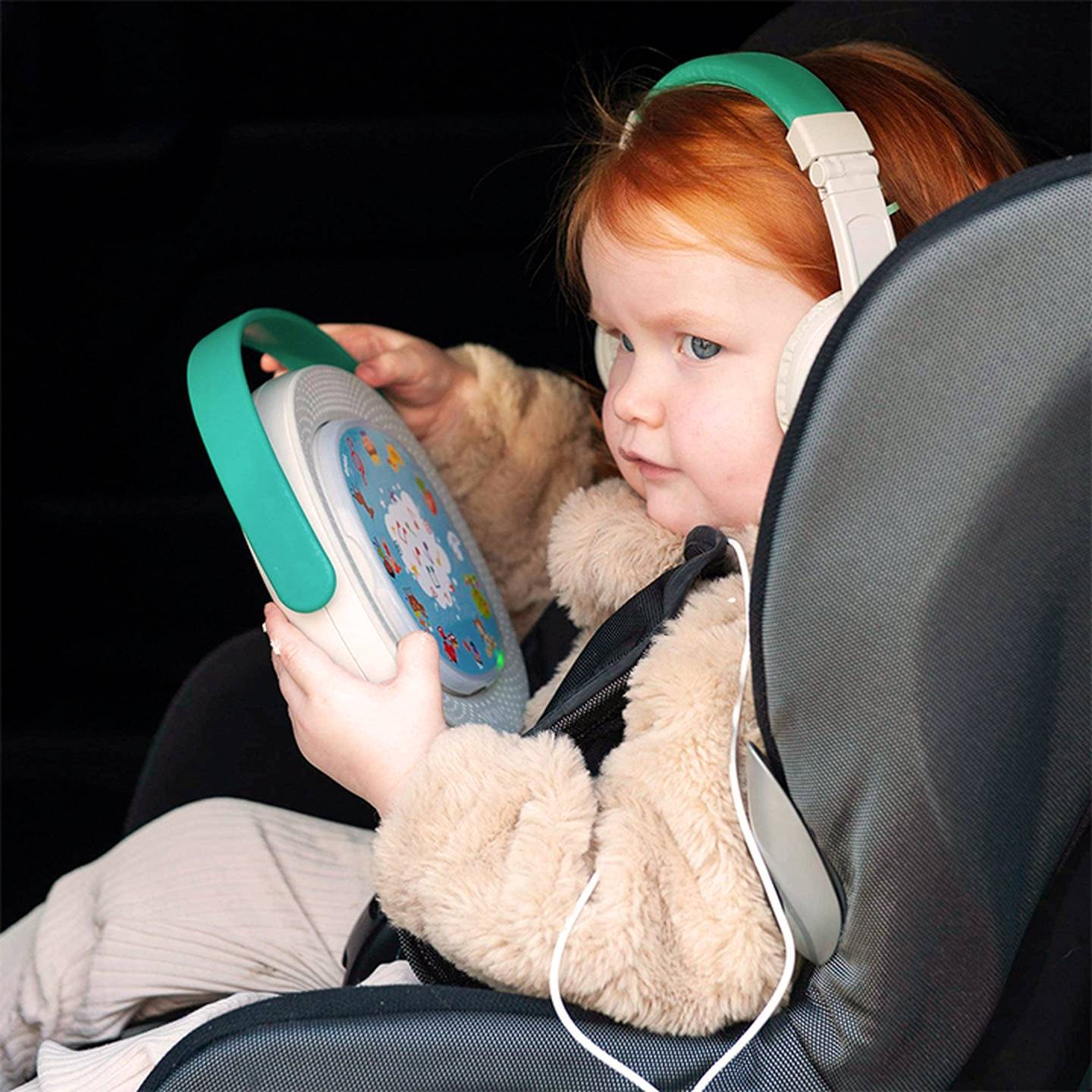 Kleines Kind mit roten Haaren, Kopfhörern, sitzt in einem Autositz, hält ein Tablet mit einer bunten Hülle in der Hand und schaut aufmerksam auf den Bildschirm.