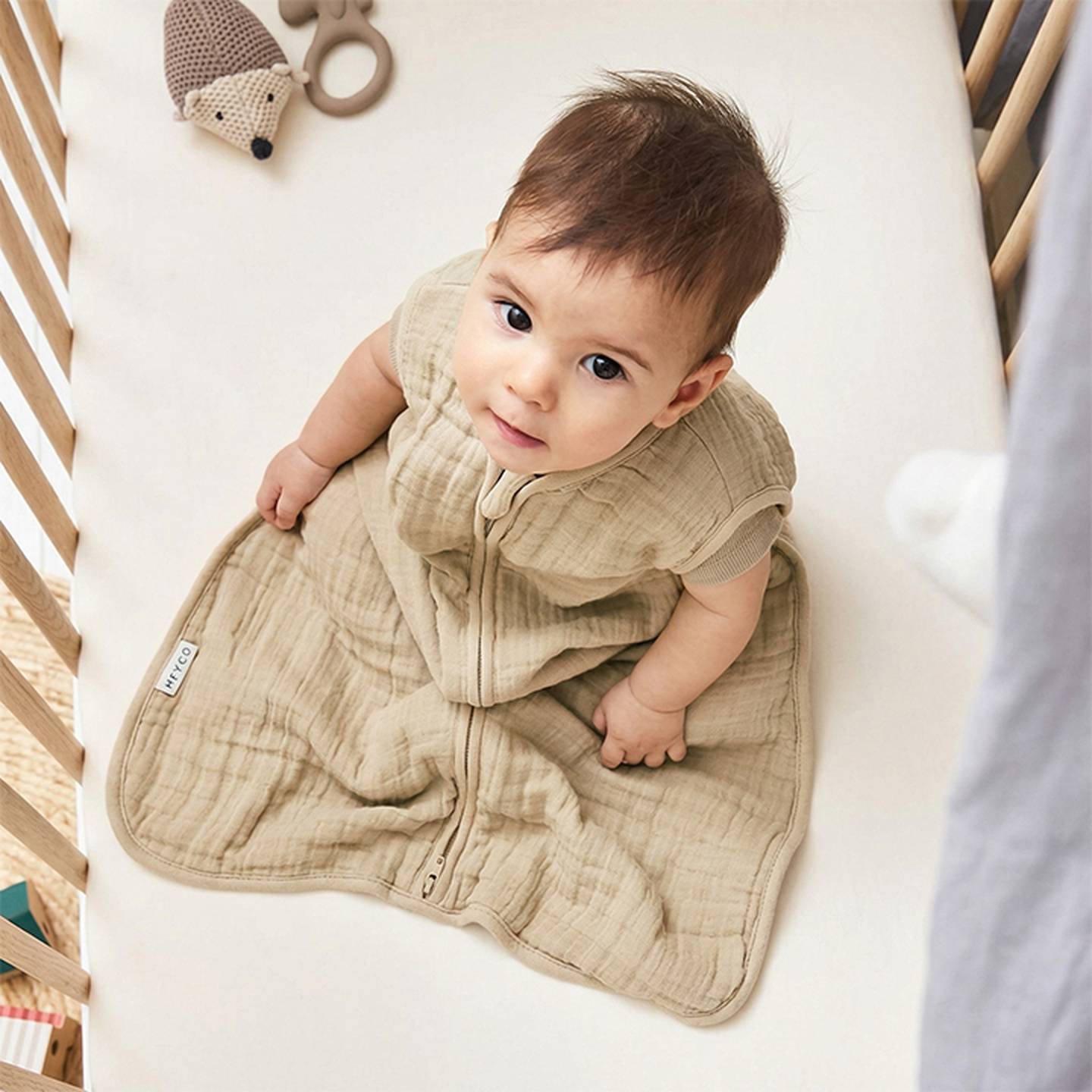 Ein Baby in einem beigen Schlafsack sitzt in einem Kinderbett und schaut nach oben. Im Hintergrund liegt auf der Matratze ein Stoffigel.
