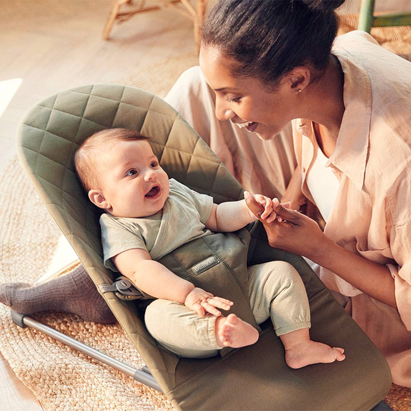 Eine Frau lächelt und hält die Hand eines Babys, das in einer grünen Babywippe auf einem gewebten Teppich sitzt, in einem warm beleuchteten Raum.