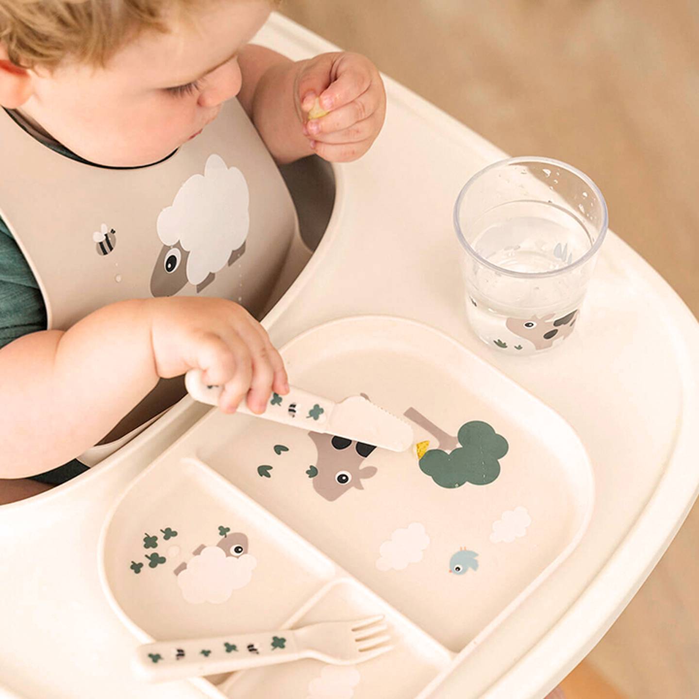 Baby im Hochstuhl mit einem Schaf-Lätzchen, isst mit einem Löffel von einem unterteilten Teller mit Tier-/Waldmotiven und trinkt aus einem Becher Wasser.