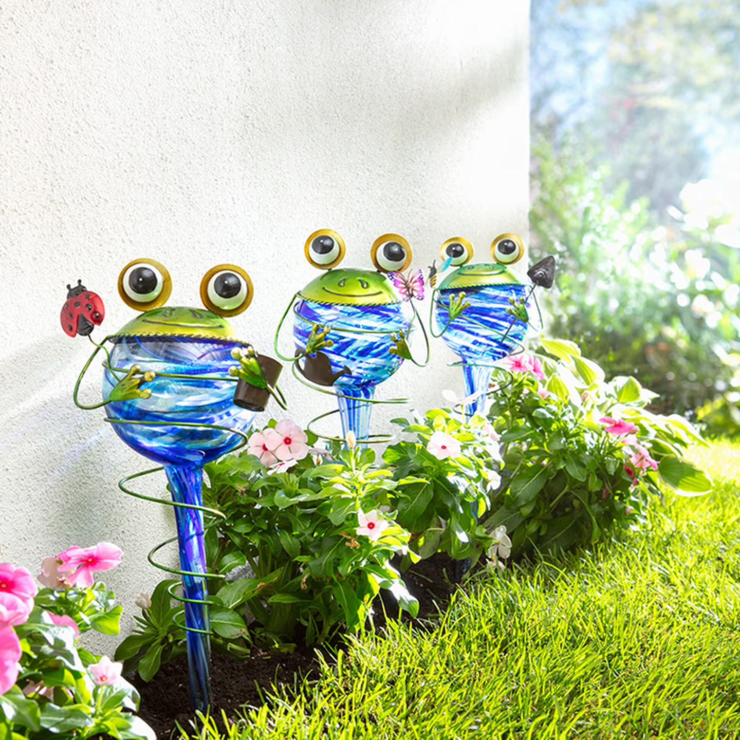 Drei verspielte Glasfrösche an Stäben für den Garten – mit großen Augen und farbenfrohen Akzenten, umgeben von leuchtend rosa Blüten und grünem Gras.