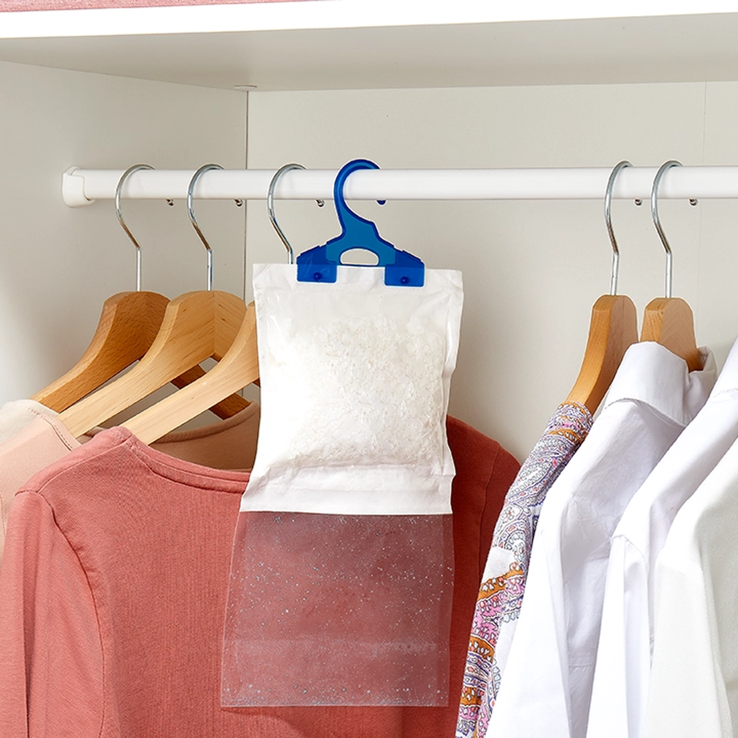 Feuchtigkeitsabsorber, der in einem Schrank zwischen Kleidungsstücken auf Holzbügeln hängt, darunter ein rosa Oberteil und ein gemustertes Hemd.
