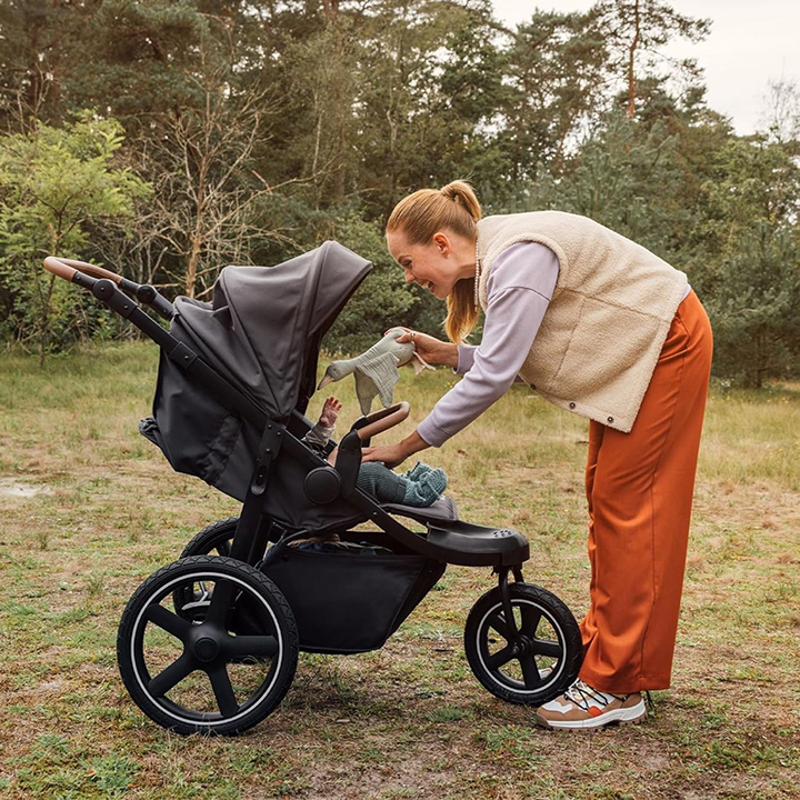 Eine Frau in einer Weste beugt sich in einer Rasenfläche über einen Kinderwagen und lächelt ein Baby an, während sie eine kleine Decke hält.
