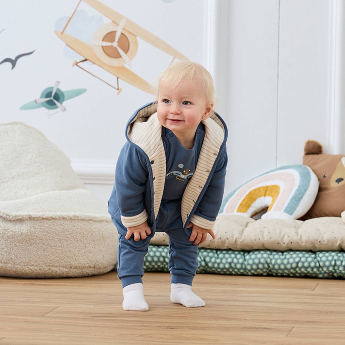 Ein Kleinkind in einem blauen Outfit steht lächelnd auf einem Holzboden; im Hintergrund sind Wanddekorationen mit Flugzeugmotiven und bunte Kissen zu sehen.