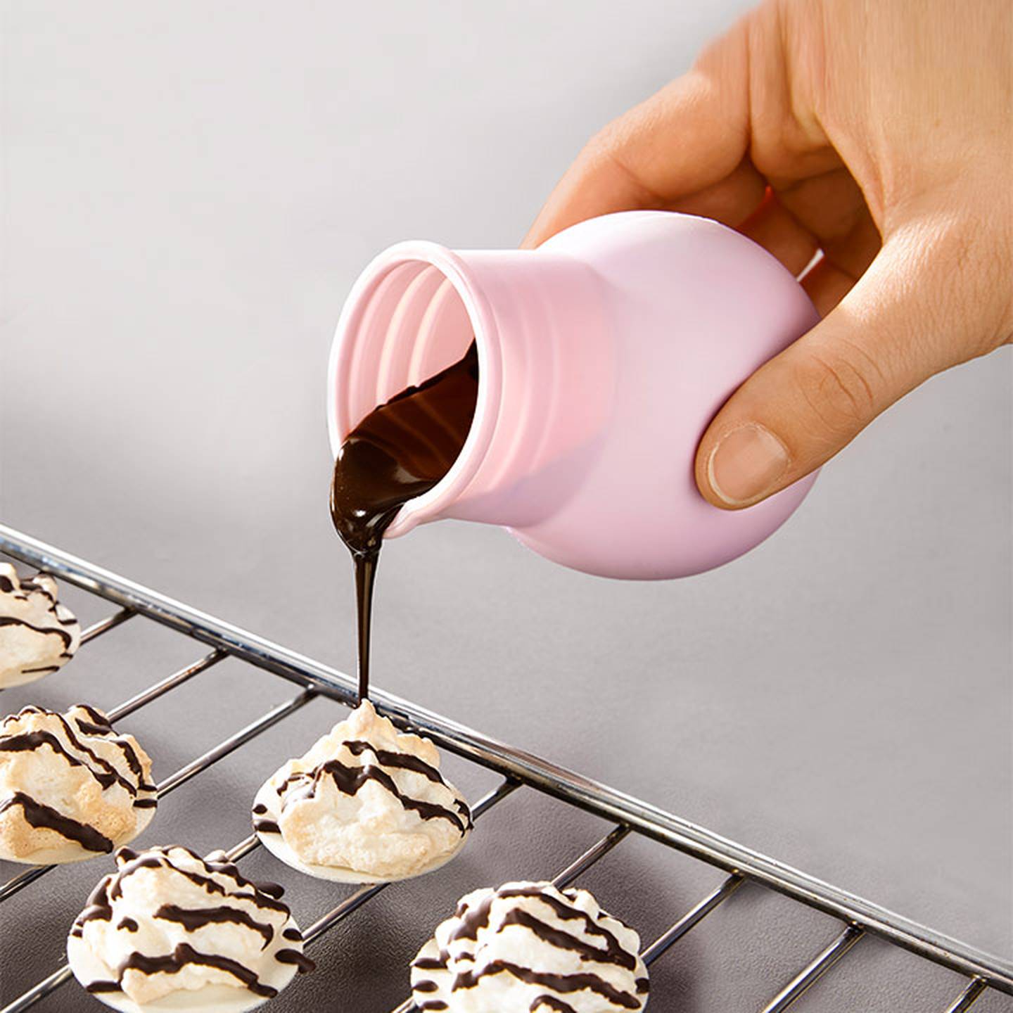 Schokoladensoße wird von Hand aus einem rosa Behälter auf Kekse auf einem Kuchengitter gegossen, wodurch Streifenmuster entstehen.