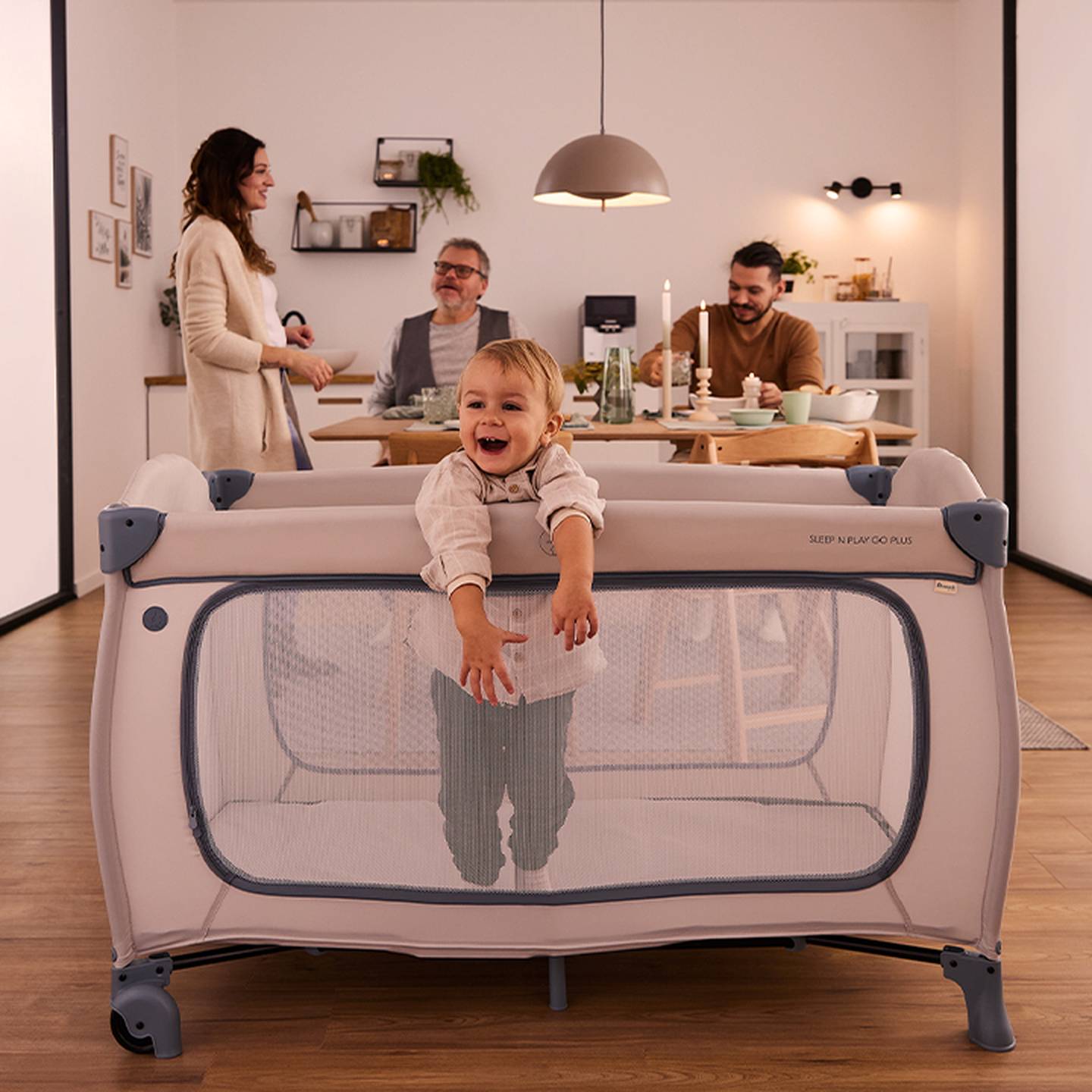 Ein fröhliches Kleinkind steht in einem Laufstall, während drei Erwachsene in einem warm beleuchteten, gemütlichen Esszimmer eine Mahlzeit an einem Tisch genießen.