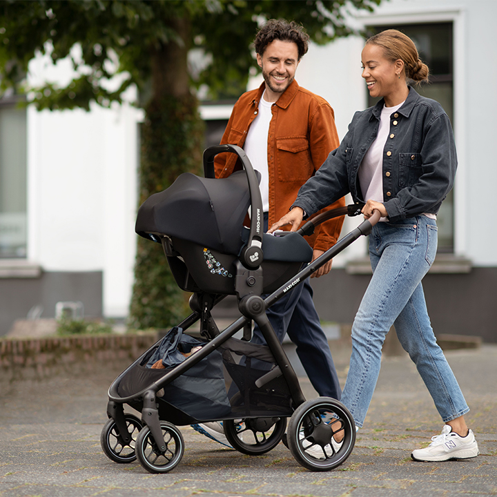 Ein lächelndes Paar spaziert draußen entlang, die Frau schiebt einen Kinderwagen, beide sind lässig in Jacken und Jeans gekleidet.