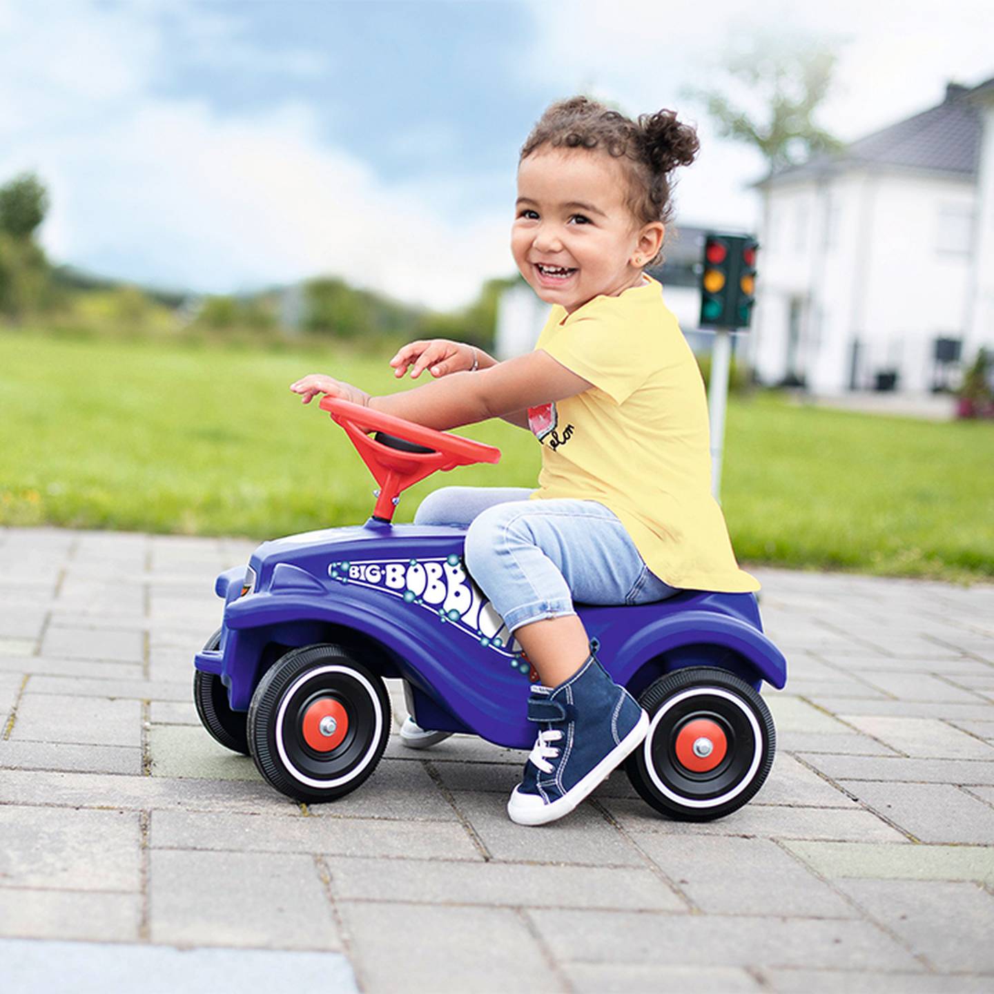 Ein lächelndes Kind fährt in einem lila Spielzeugauto mit rotem Lenkrad, gekleidet in einem gelben Hemd und Jeans, auf einem Bürgersteig neben einer Rasenfläche.
