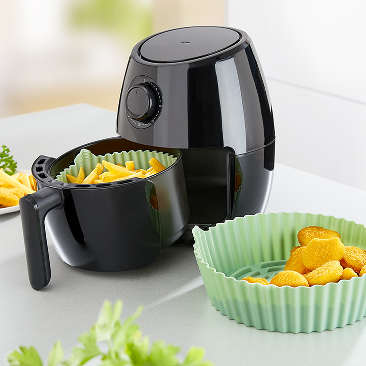 Eine schwarze Heißluftfritteuse auf einer Arbeitsplatte, mit Silikon-Backeinlagen darin und daneben, die Pommes frites und Nuggets enthalten.
