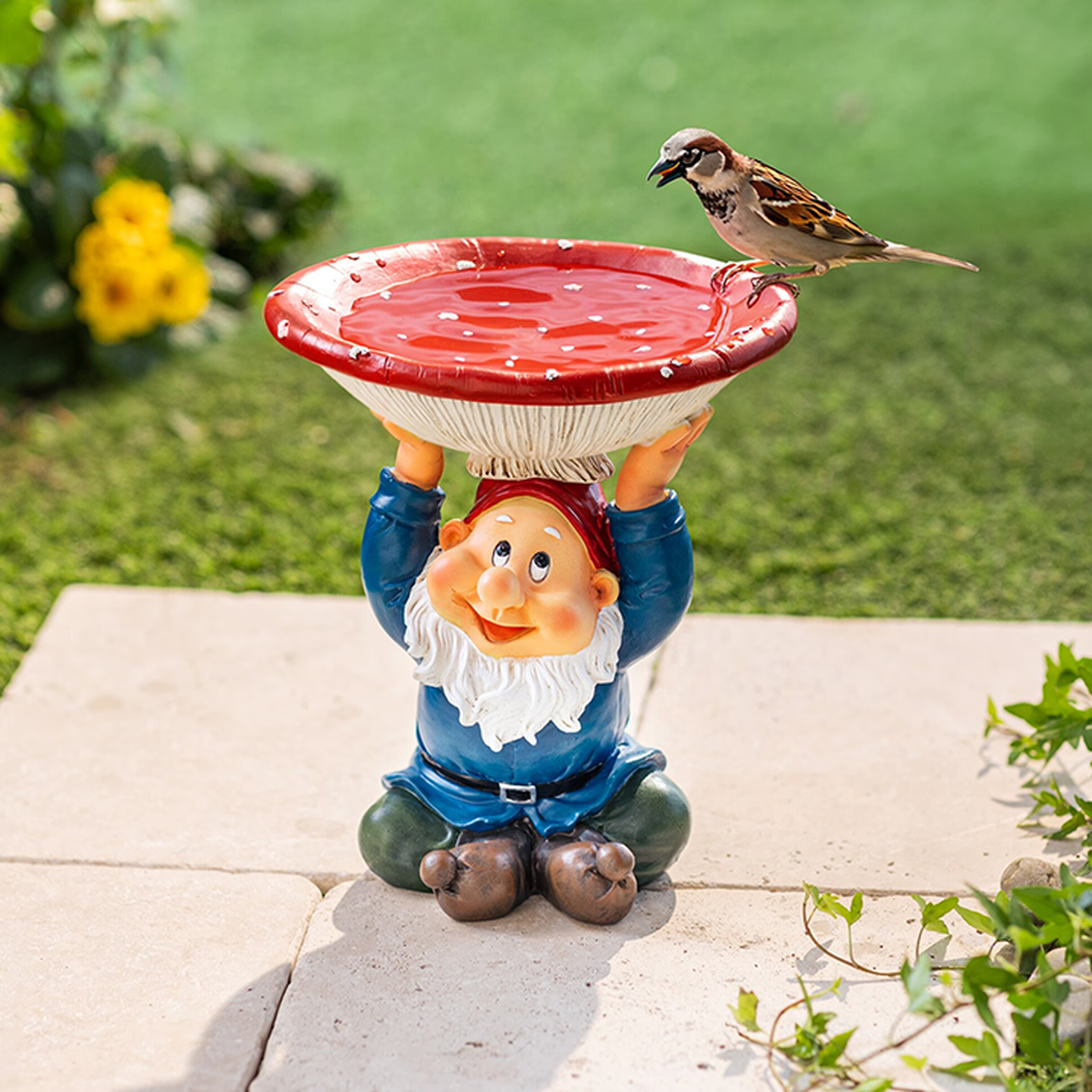 Ein Gartenzwerg, der ein rotes, pilzförmiges Vogelbad hochhält, auf dessen Rand ein Spatz sitzt – inmitten eines üppig grünen Gartens.