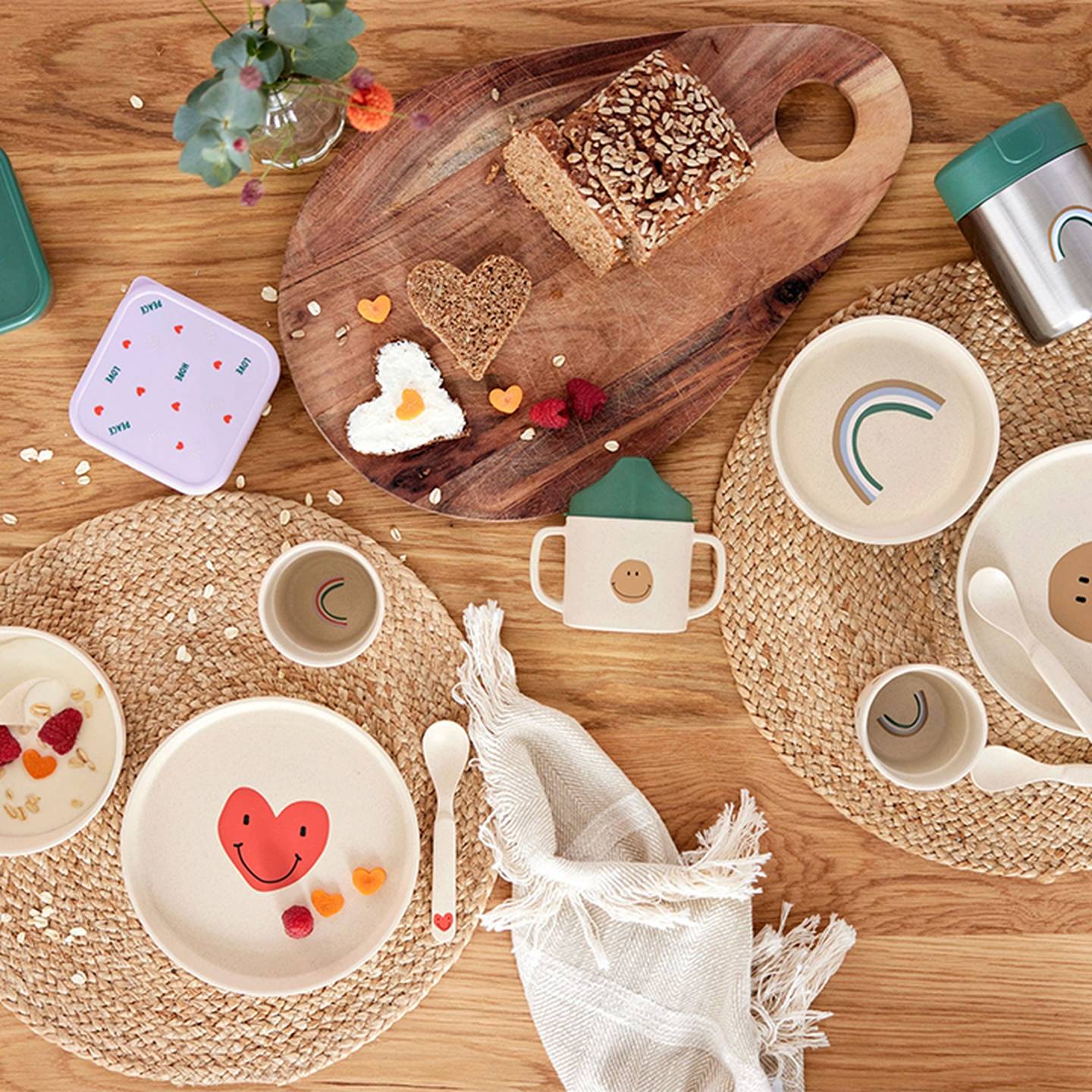 Ein gedeckter Tisch mit verspieltem Kindergeschirr, auf dem lächelnde Gesichter und Regenbögen abgebildet sind, daneben Brot, Obst und eine kleine Vase mit Blumen.