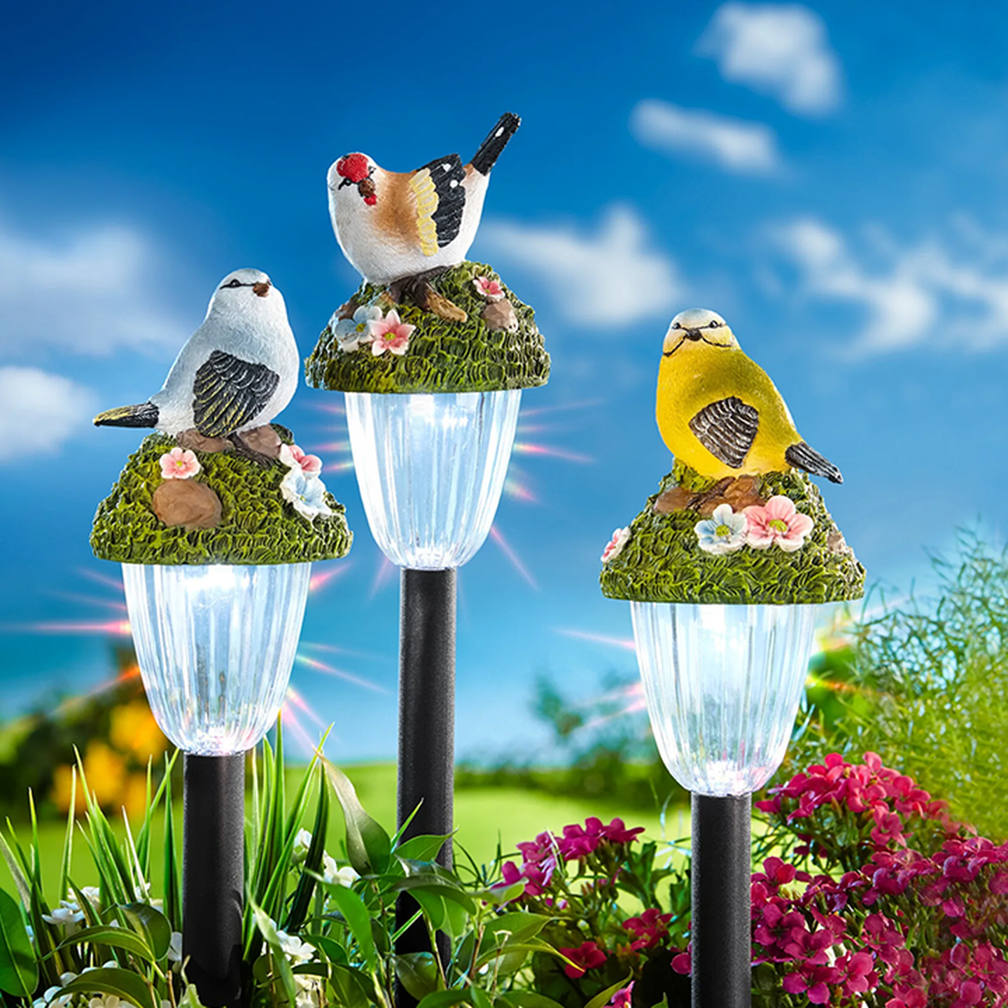 Drei Solar-Gartenleuchten, gekrönt von dekorativen Vogelfiguren und Blumen, vor einem klaren blauen Himmel mit sattem Grün und rosa Blüten.