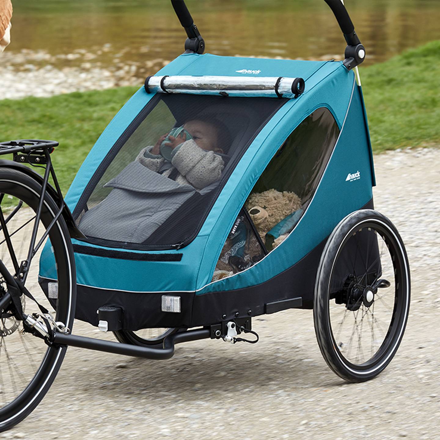 Ein Baby in einem blauen Fahrradanhänger mit großem Fenster hält eine Flasche. Ein Teddybär steht daneben. Der Anhänger ist an einem Fahrrad befestigt.