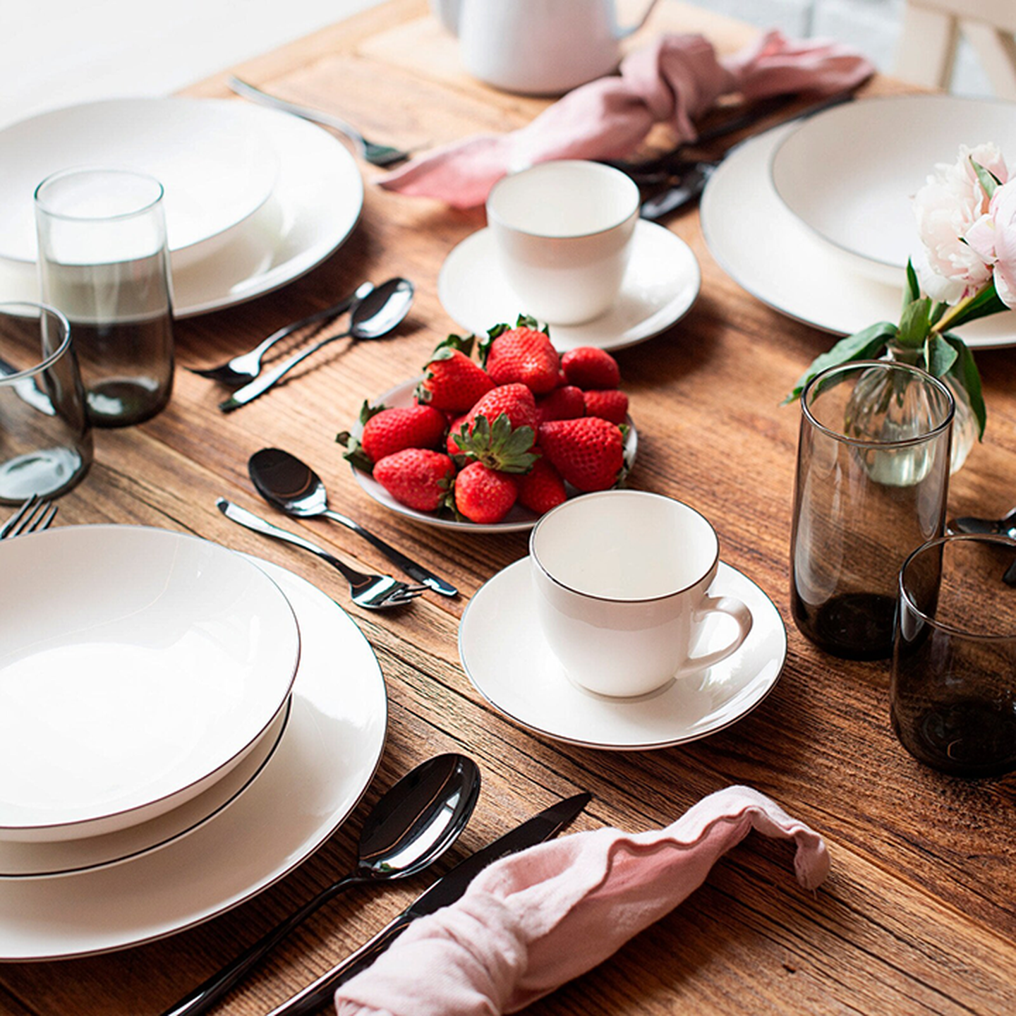 Eine elegante Tischdekoration mit weißem Geschirr und Tassen, frischen Erdbeeren, Besteck, Gläsern, rosa Servietten und einem floralen Tischgesteck auf einem Holztisch.