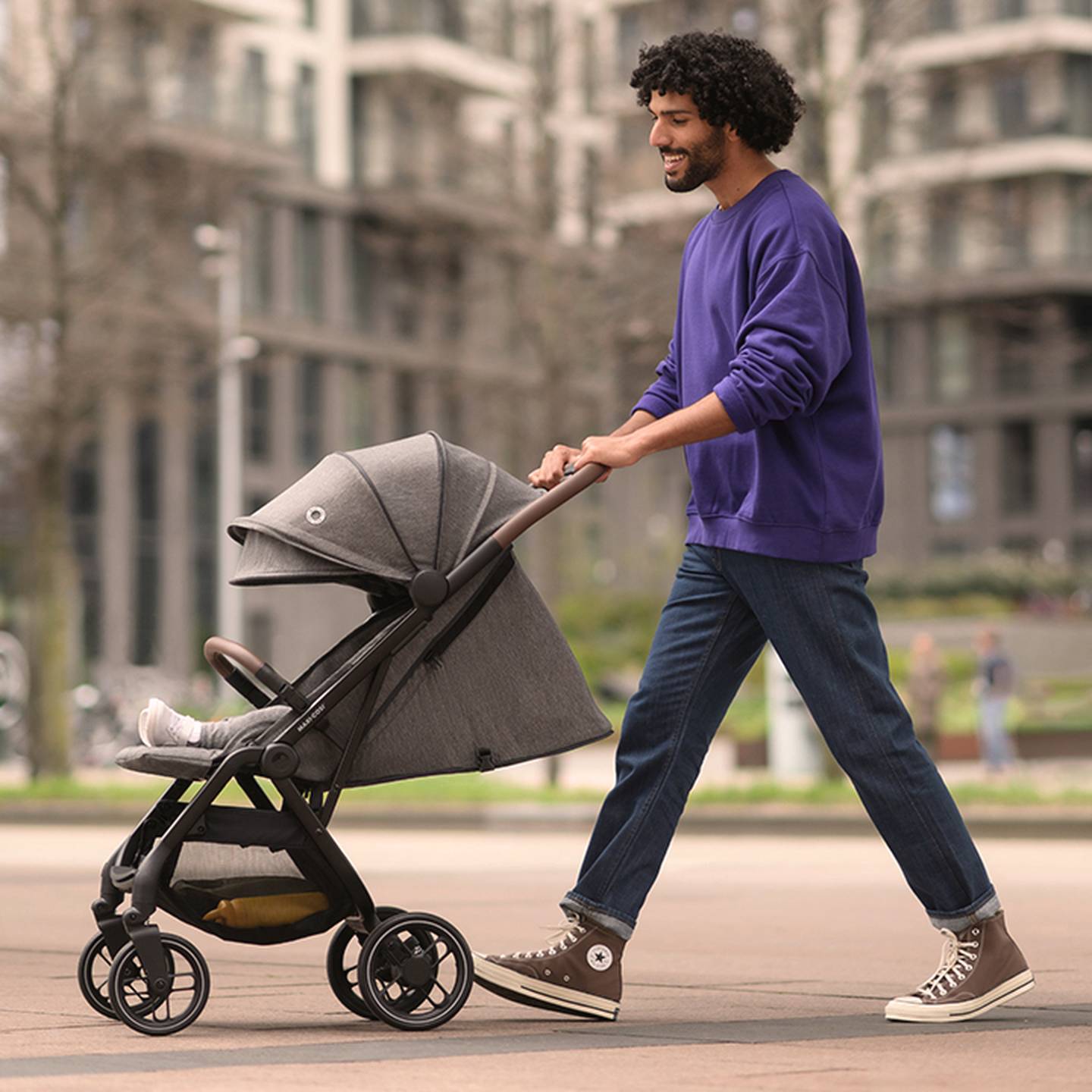 Ein Mann in einem lila Pullover schiebt einen Kinderwagen auf einem Bürgersteig. Er trägt Jeans und braune Turnschuhe. Im Hintergrund sind Stadtgebäude zu sehen.