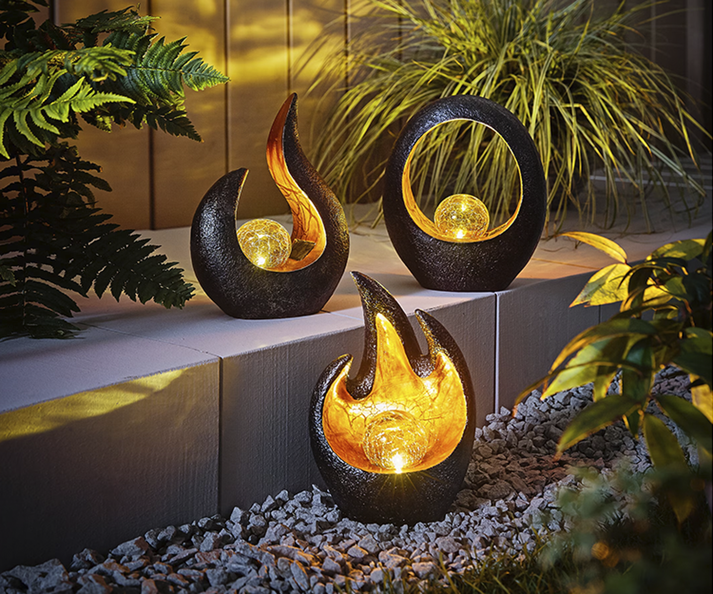 Drei dekorative Gartenskulpturen mit leuchtenden Kugeln, eingebettet zwischen Grünpflanzen und Steinen. Jede Skulptur hat eine einzigartige Form und strahlt ein warmes Licht aus.