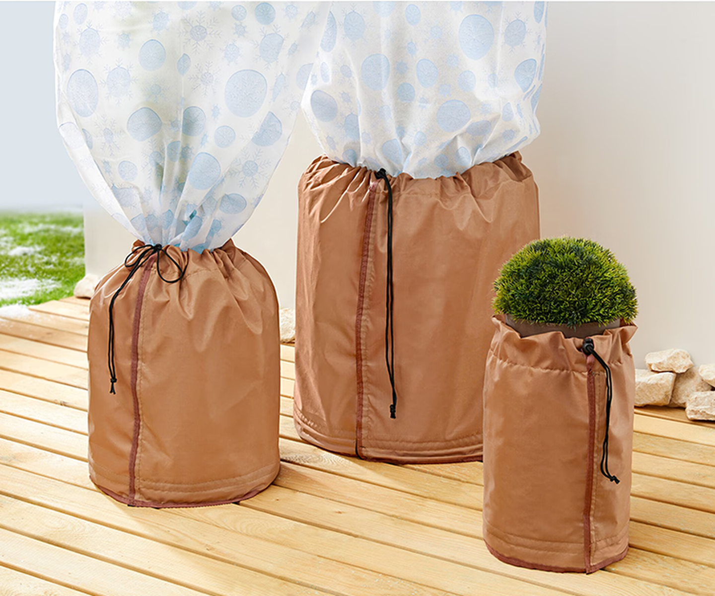 Drei Pflanzen auf einer Holzterrasse, bedeckt mit braunen Schutzbeuteln. Ein Beutel ist teilweise geöffnet und gibt den Blick auf einen kleinen grünen Strauch im Inneren frei.