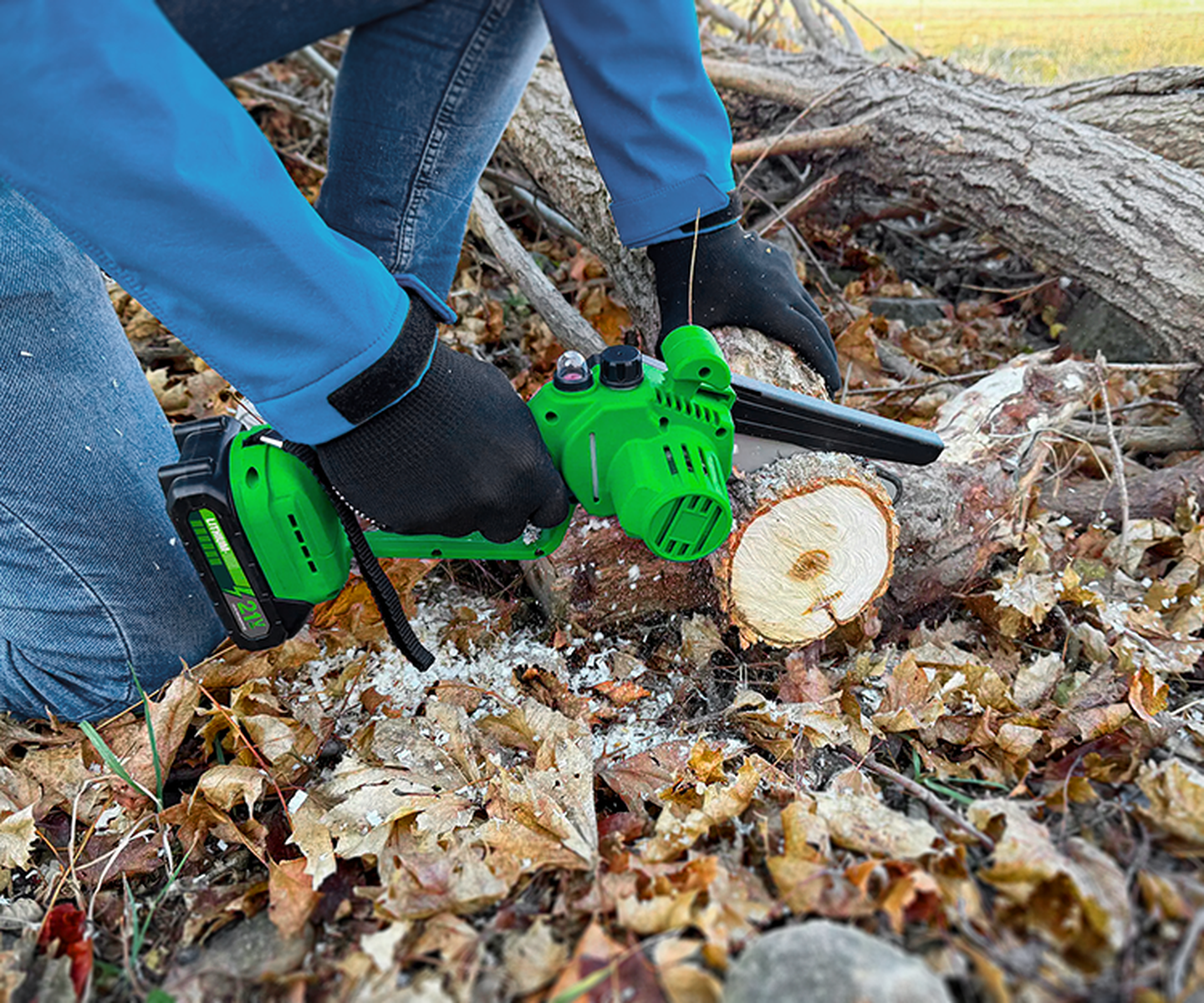 Person in blauer Jacke, die mit einer grünen Elektrokettensäge einen heruntergefallenen Ast zwischen verstreuten Blättern zersägt.