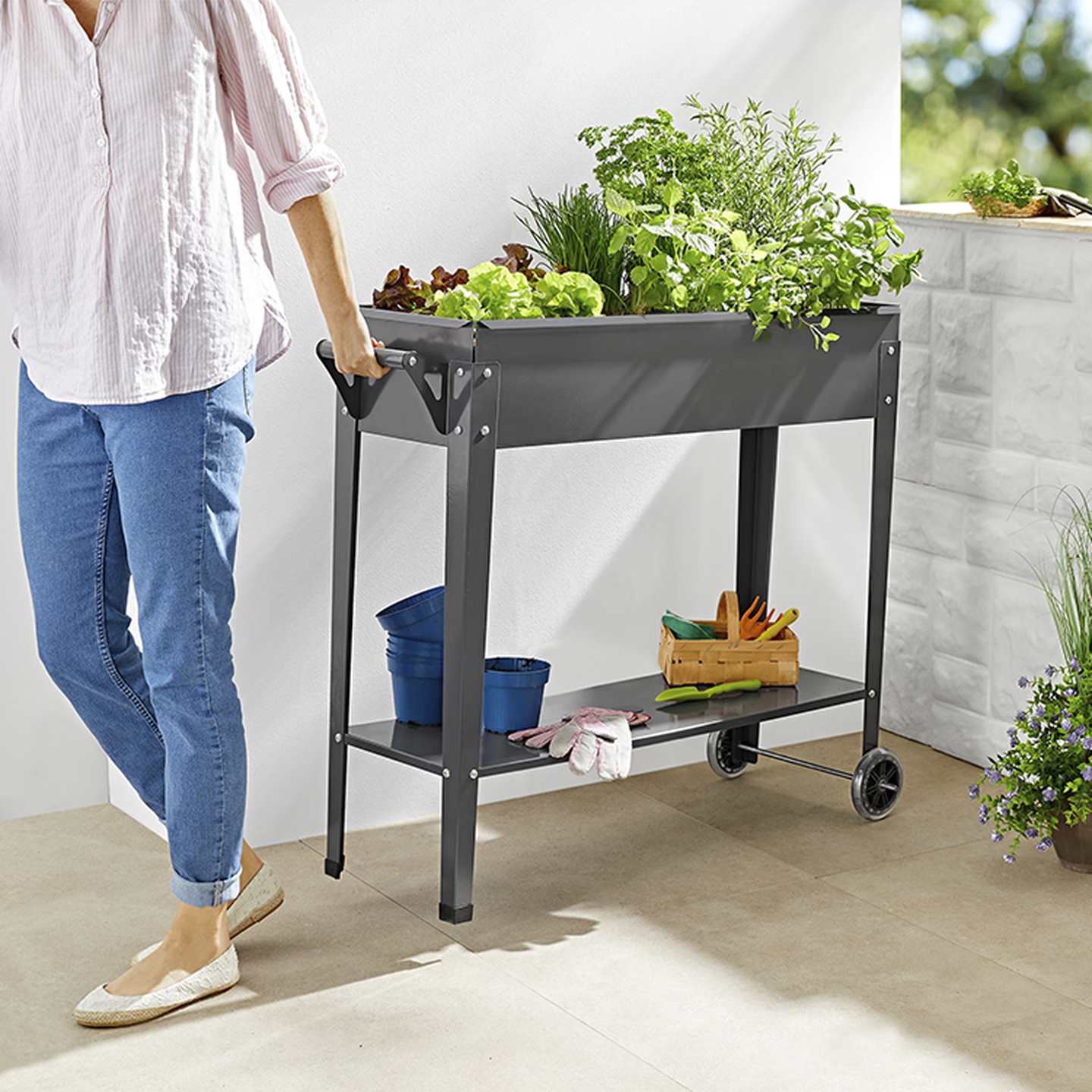 Person in Bluejeans und rosa Hemd, die ein erhöhtes Gartenbeet auf Rädern pflegt, mit einer Vielzahl von Grünpflanzen und Gartengeräten in der Nähe.