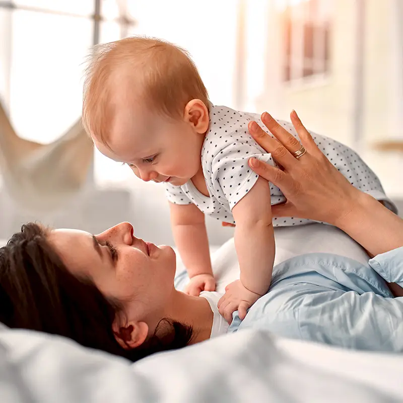 Eine Person liegt auf einem Bett und lächelt ein Baby in einem gepunkteten Outfit an, das spielerisch auf ihrer Brust in einem sonnenbeschienenen Raum liegt.