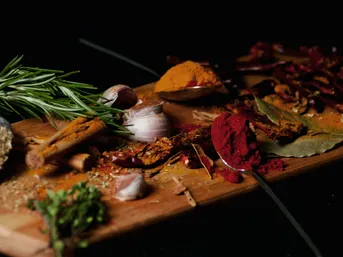 Ein Holzbrett mit verschiedenen Gewürzen: Kurkuma, Paprika, Lorbeerblätter, Rosmarin, Knoblauch und mehr, angeordnet mit Löffeln auf einem dunklen Hintergrund.