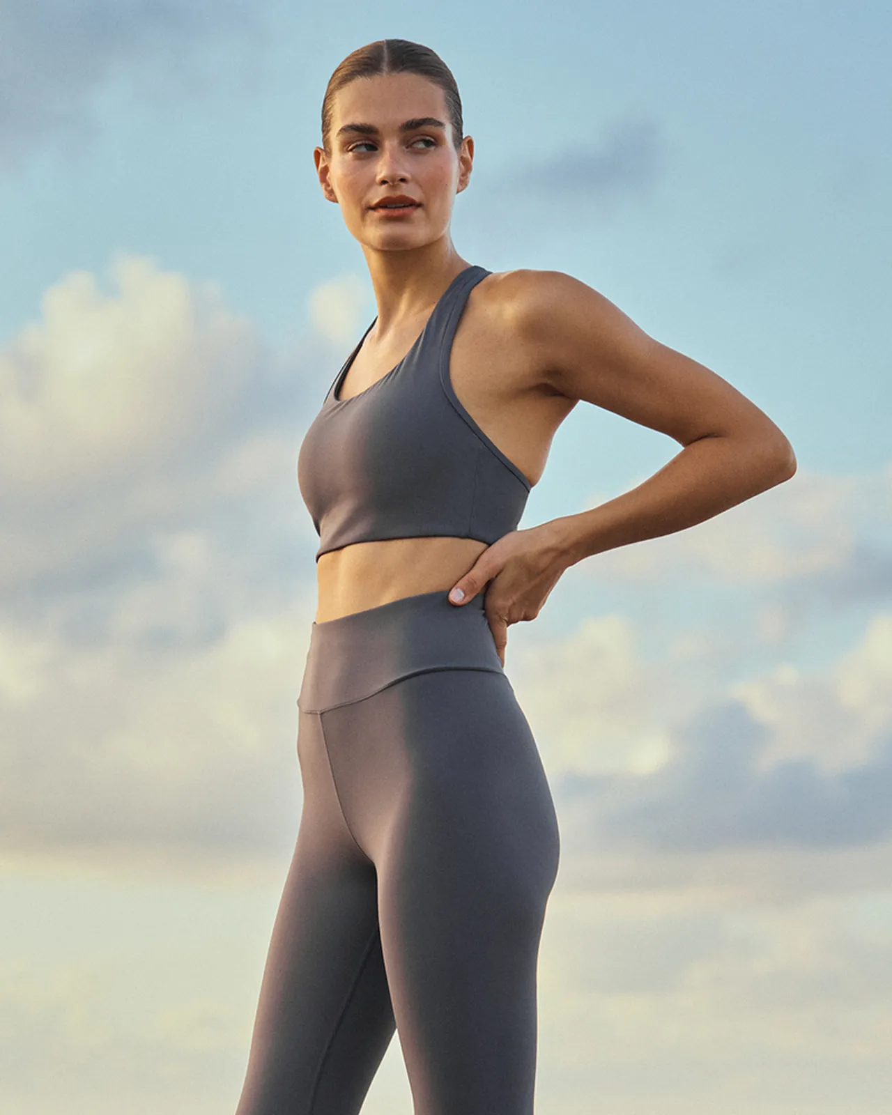 Woman in gray sports bra and high-waist leggings standing outdoors with hand on hip, looking to the side against a cloudy sky.