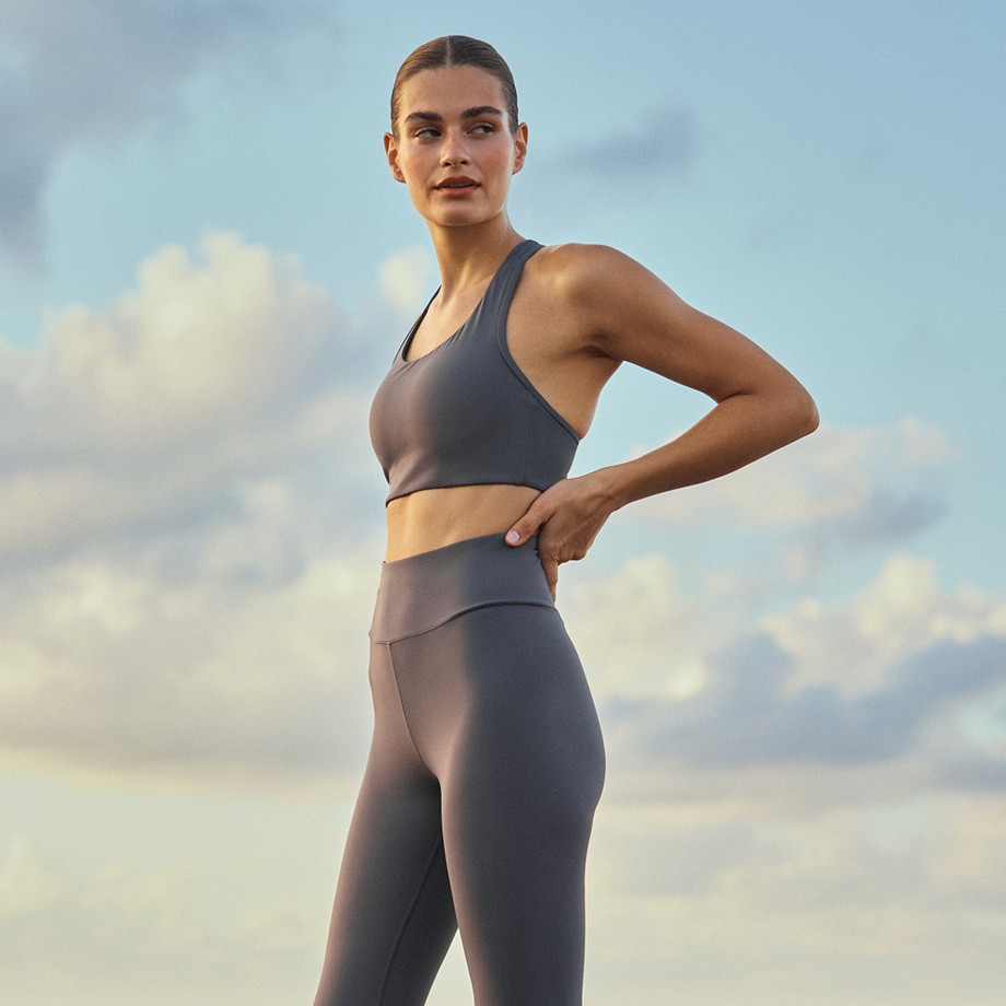 Woman in gray sports bra and high-waist leggings standing outdoors with hand on hip, looking to the side against a cloudy sky.