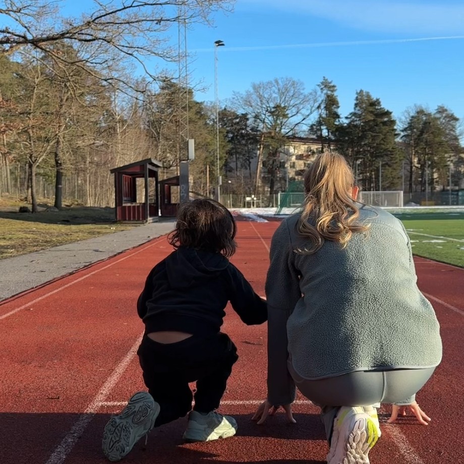 Child and adult crouching at a track starting line in a park, surrounded by trees and a clear blue sky.