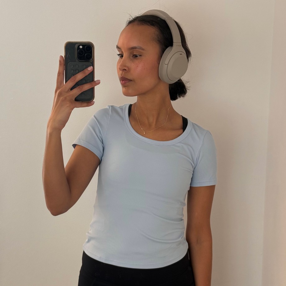 Person in a light blue shirt and headphones taking a mirror selfie with a smartphone against a plain wall.