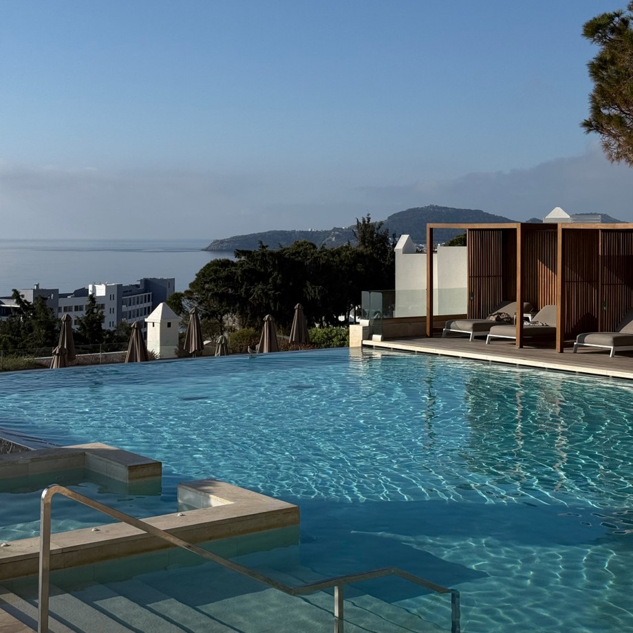 A serene infinity pool with wooden cabanas overlooking a calm sea and distant hills under a clear blue sky.