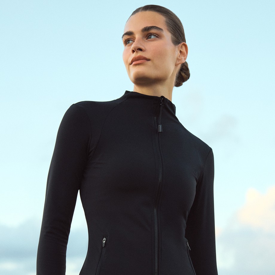 Woman in a fitted black zip-up jacket with hair in a bun, gazing upward against a pale blue sky.