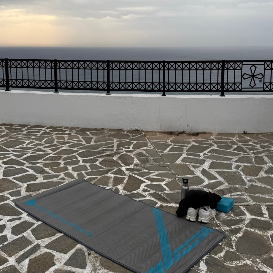 Yoga mat, shoes, and water bottle on a stone patio overlooking the ocean, with a decorative black fence and cloudy sky in the background.