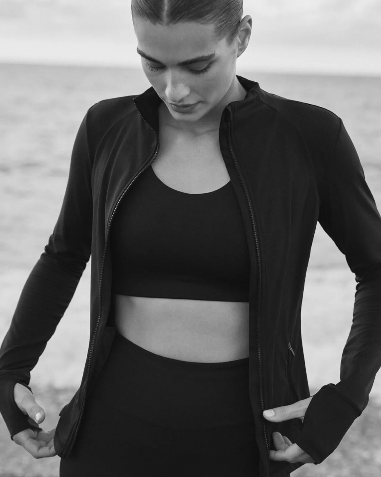 Woman in black sports bra, zip-up jacket and high-waisted leggings adjusting jacket on a calm beach, looking down.