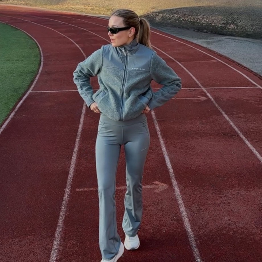 Woman in athletic wear and sunglasses standing confidently on a red running track, with her hands on her hips, in a sunny outdoor setting.