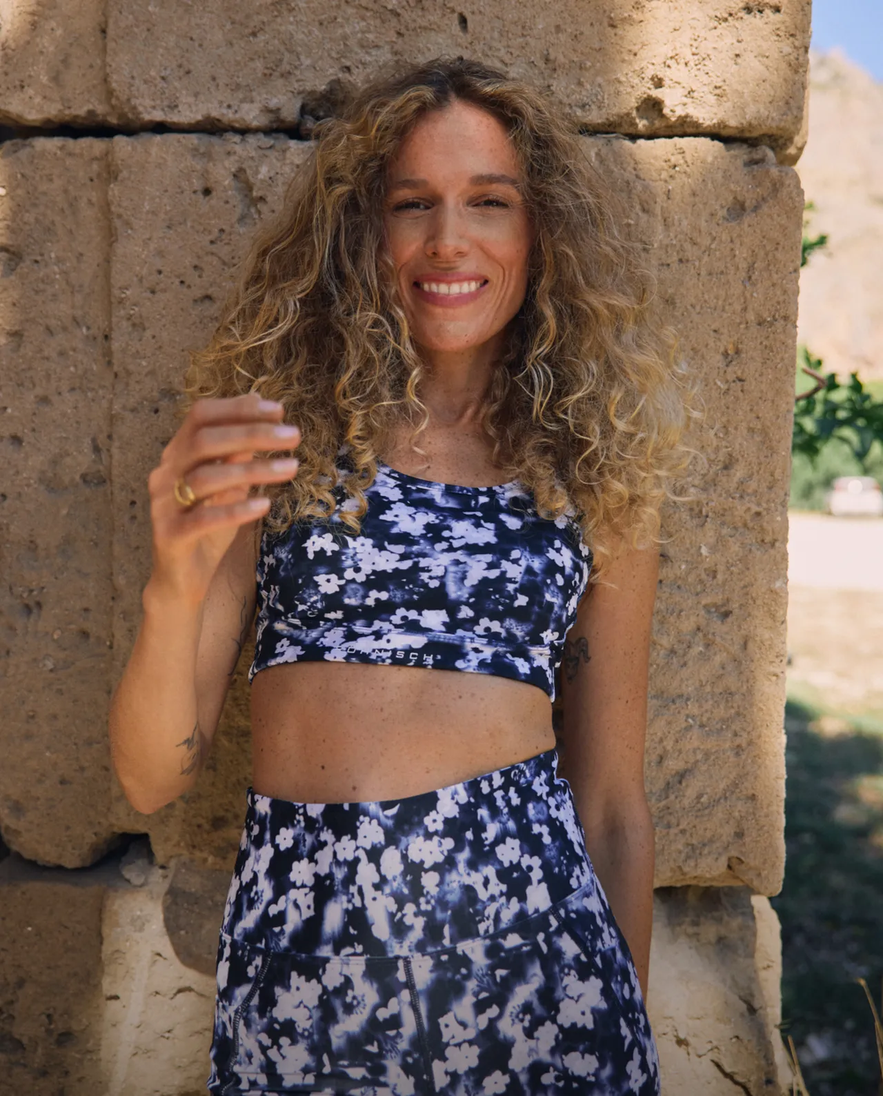 Smiling person with curly hair in floral activewear stands against a stone wall outdoors.