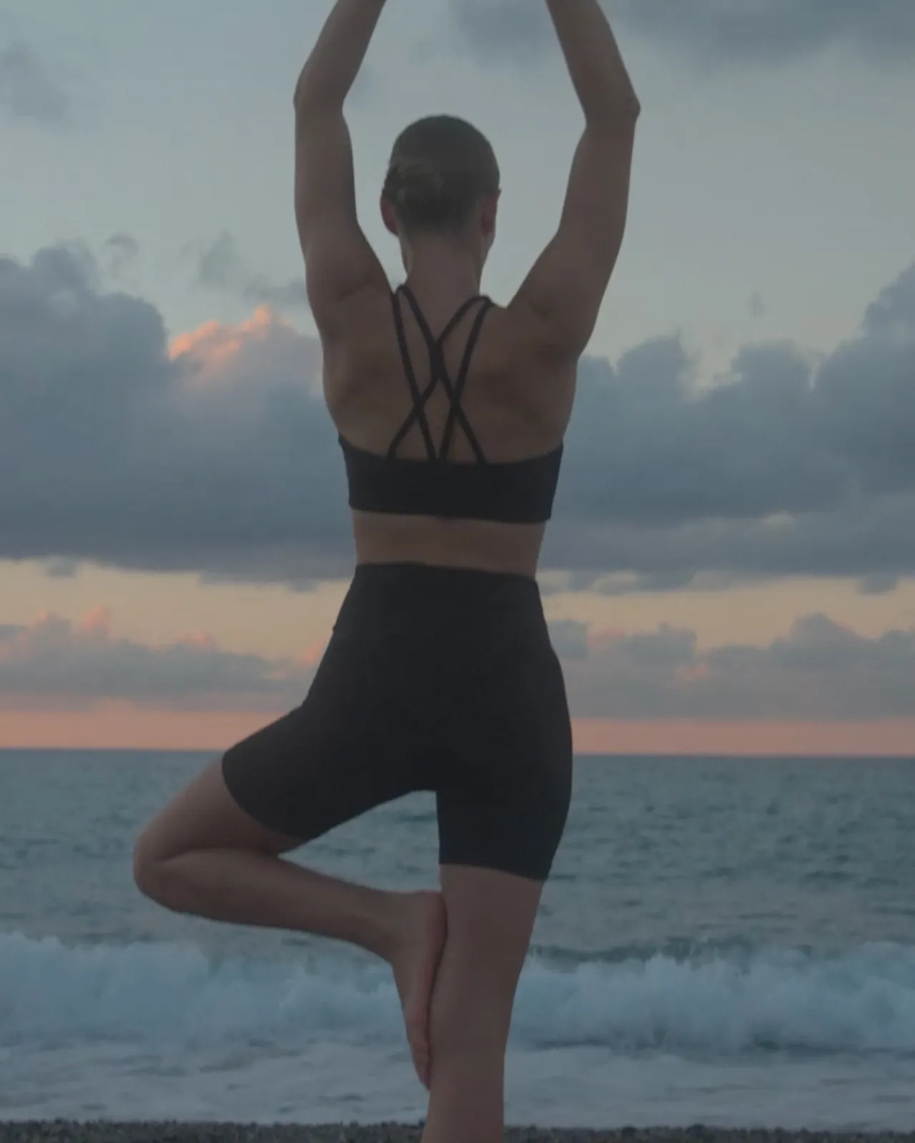Person practicing yoga in a tree pose on a beach at sunset, wearing a black sports outfit, with waves behind and a cloudy sky.