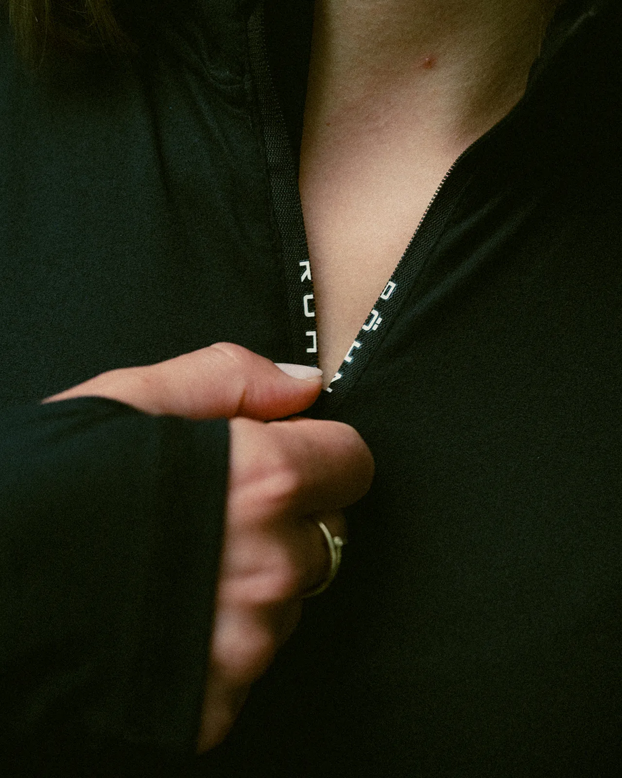 Person zipping up a black jacket, showing a partial view of their hand and the jacket's collar with text on the zipper.